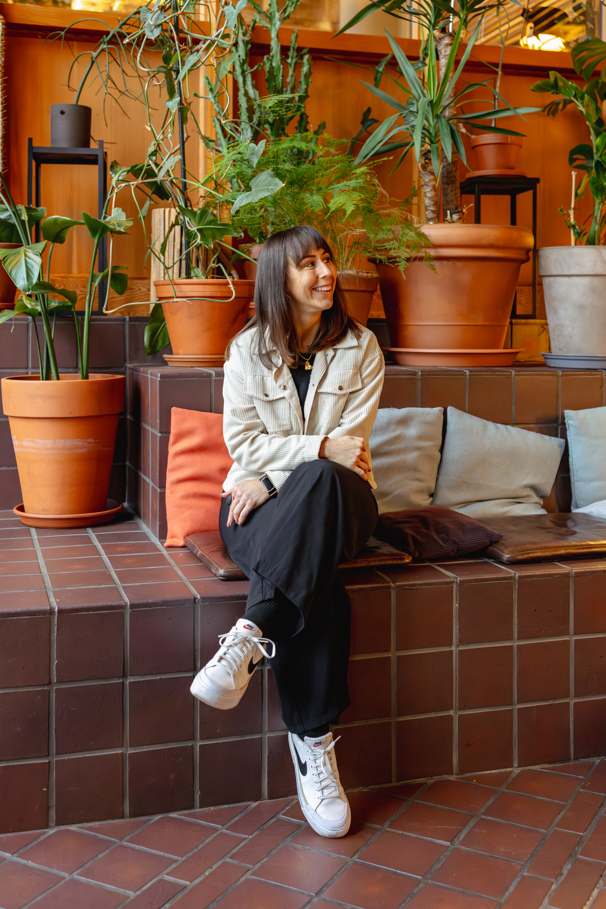 A woman sitting on a tiled bench surrounded by large potted plants, smiling and looking to her right in a cozy indoor space with wooden paneling.