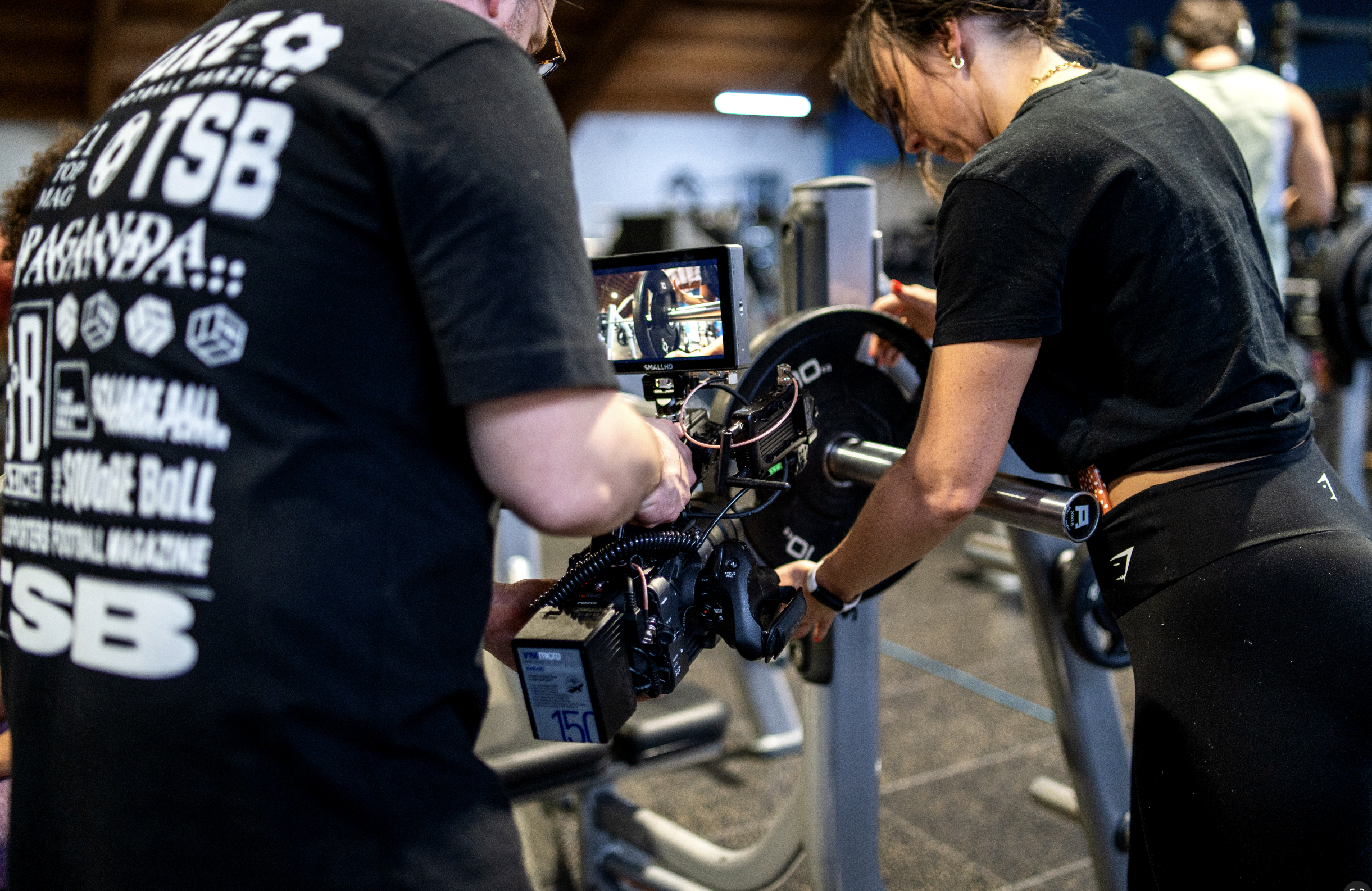 A woman adjusts a barbell on a weightlifting machine while a person films the scene with a professional camera. They are inside a gym.