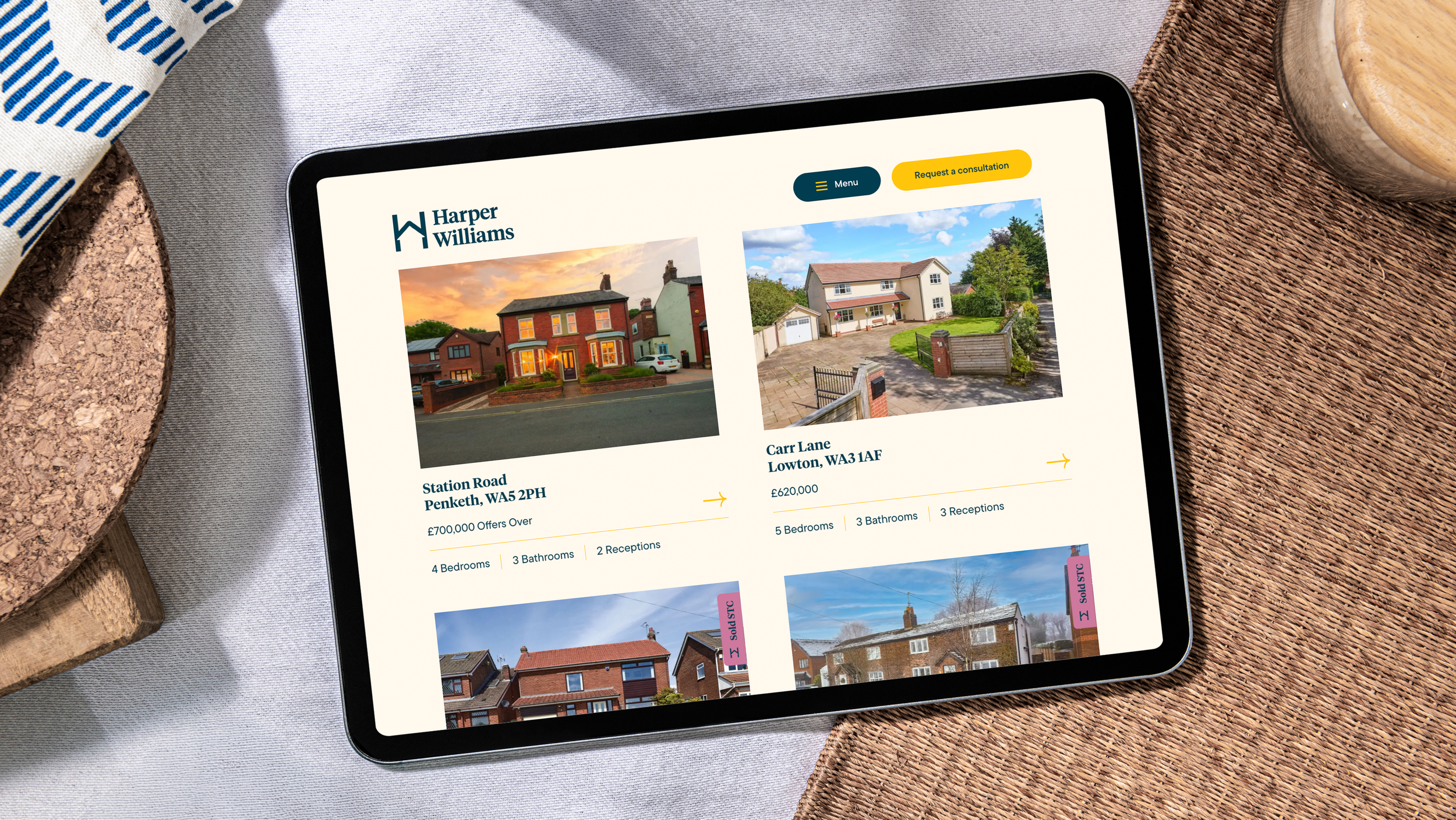 Tablet displaying a real estate website with listings and photos of houses, placed on a woven mat surrounded by decorative items.
