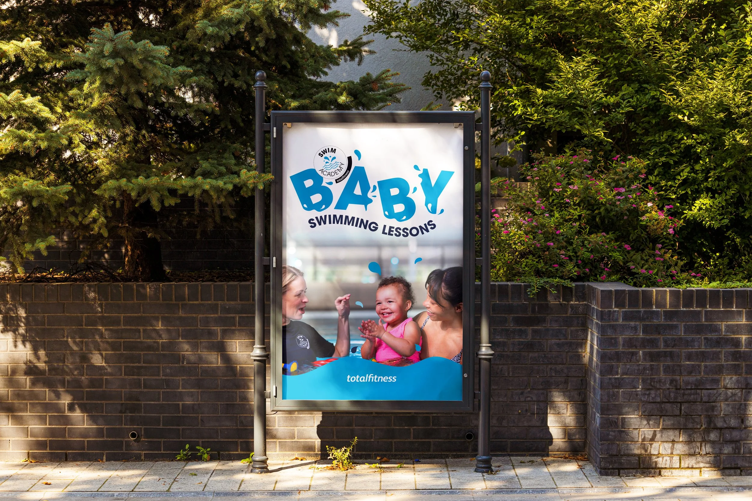 Sign advertising baby swimming lessons at Total Fitness, featuring a young girl in a pink swimsuit being held and coached by two women at a swimming pool.