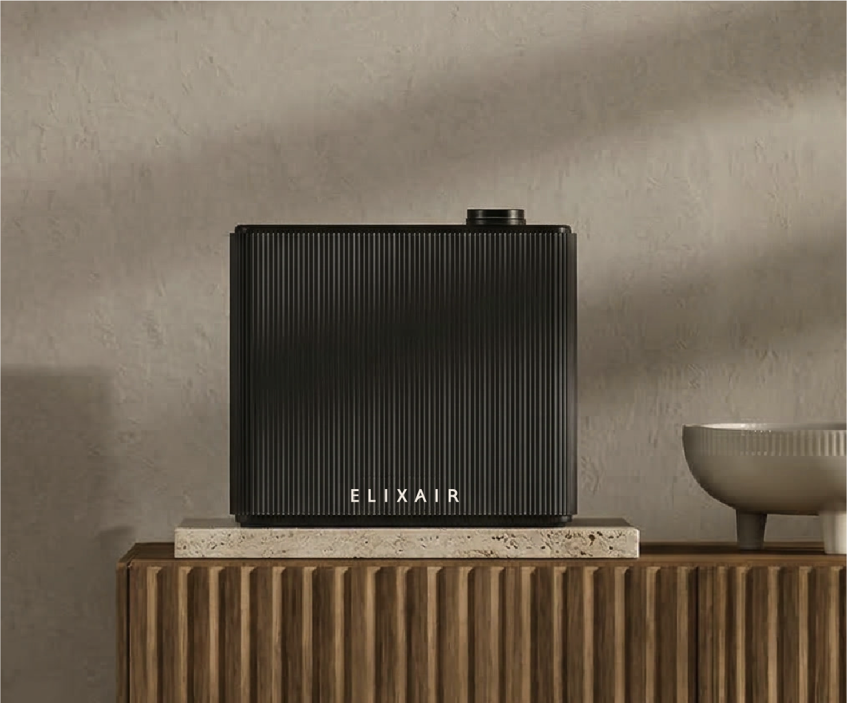 A black Elixair air purifier with a ribbed textured surface placed on a wooden surface, next to a bowl and a small white object, against a neutral wall background.