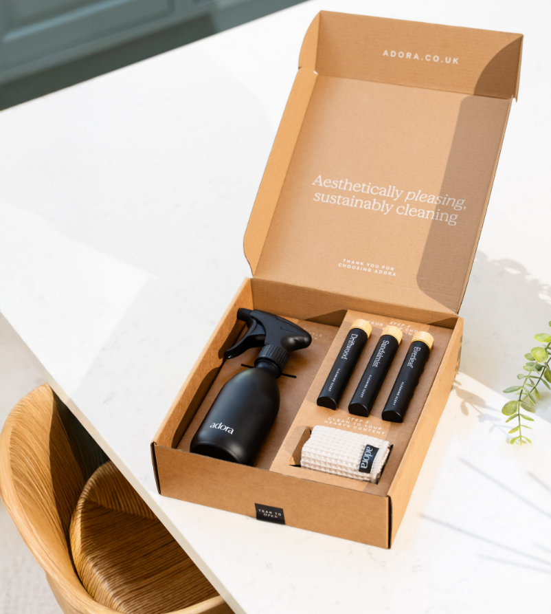 Open cardboard box from adora.co.uk containing a black spray bottle, three small tubes labeled 'Diamond,' 'Sanitizer,' and 'Dirtbreaker,' a small cloth, and a cleaning sponge on a white surface with a wooden chair nearby.