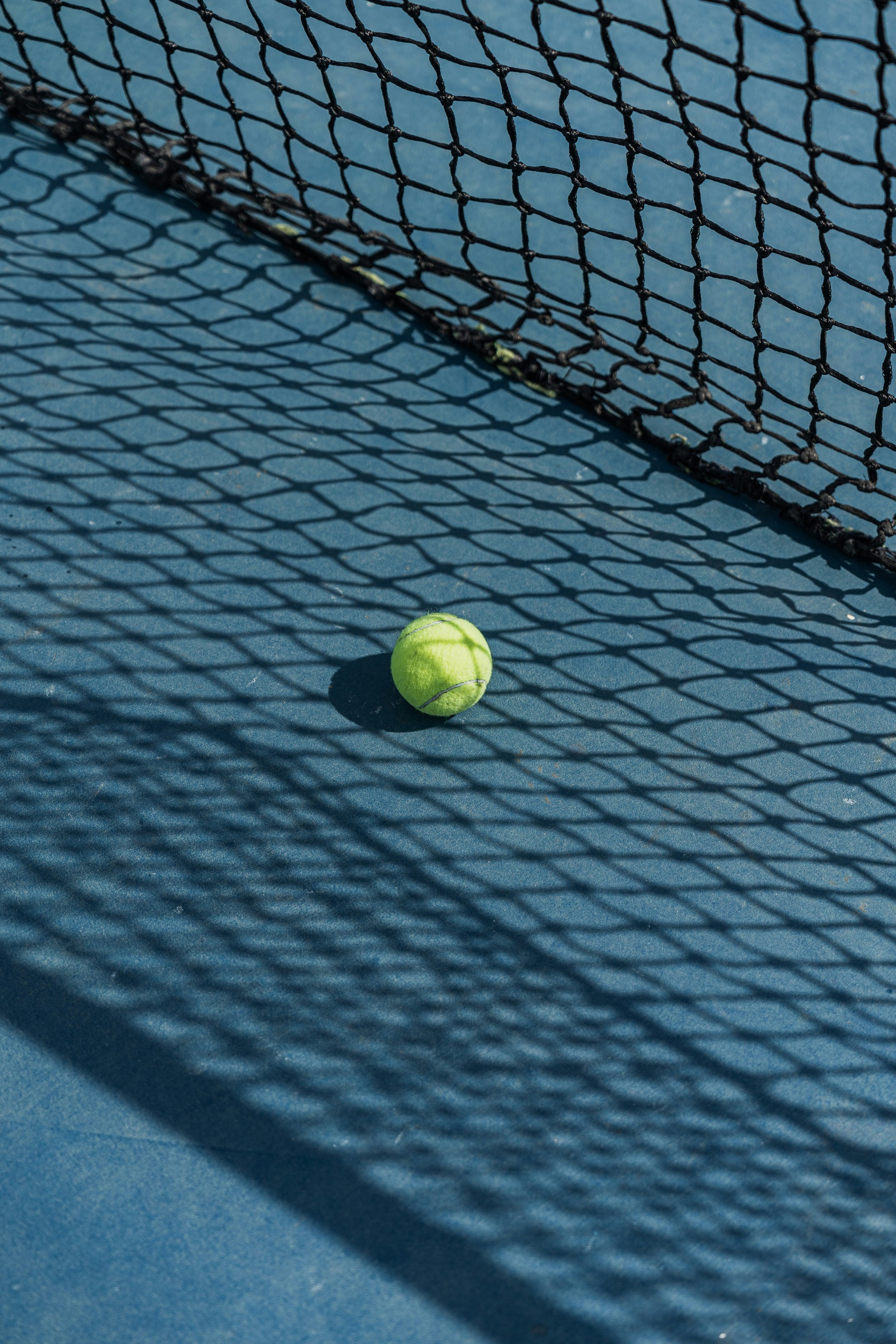 A yellow tennis ball resting on a blue tennis court, casting a shadow underneath. A tennis net is propped against the court, creating a pattern of shadow and light.