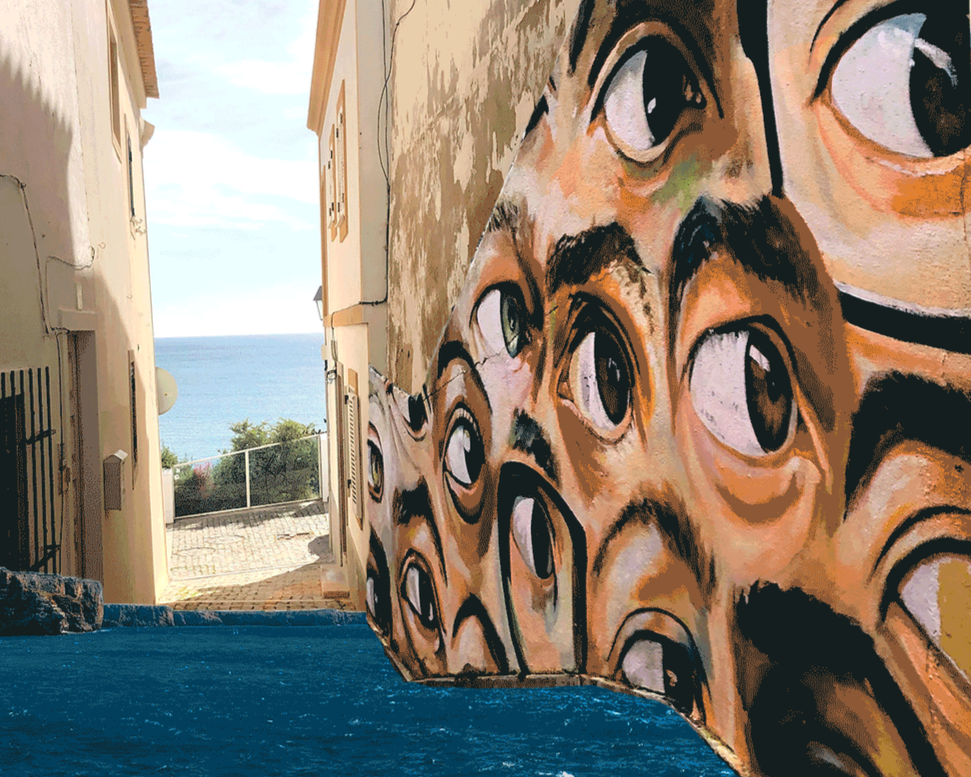 Colorful mural of multiple stylized eyes painted on a wall in a narrow alleyway, overlooking an open sea view with blue sky.
