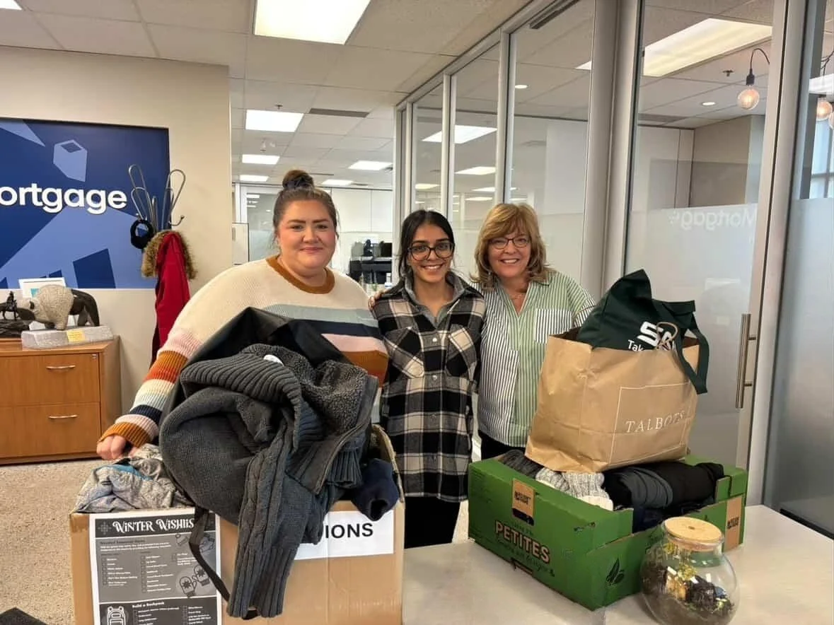 Three women standing together inside an office, smiling. They are standing behind boxes filled with clothes and bags, participating in a donation or charity event.