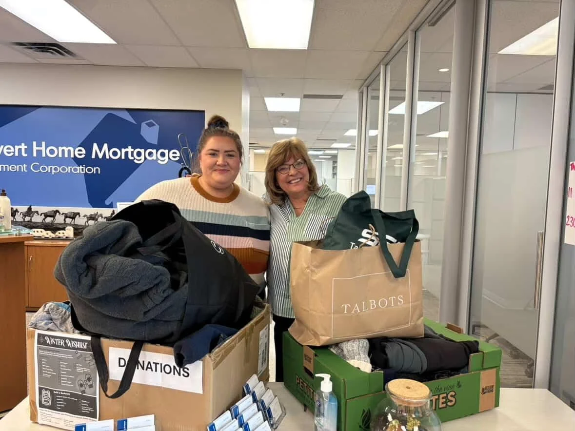Two women standing behind donation boxes filled with clothing items inside an office or community center.