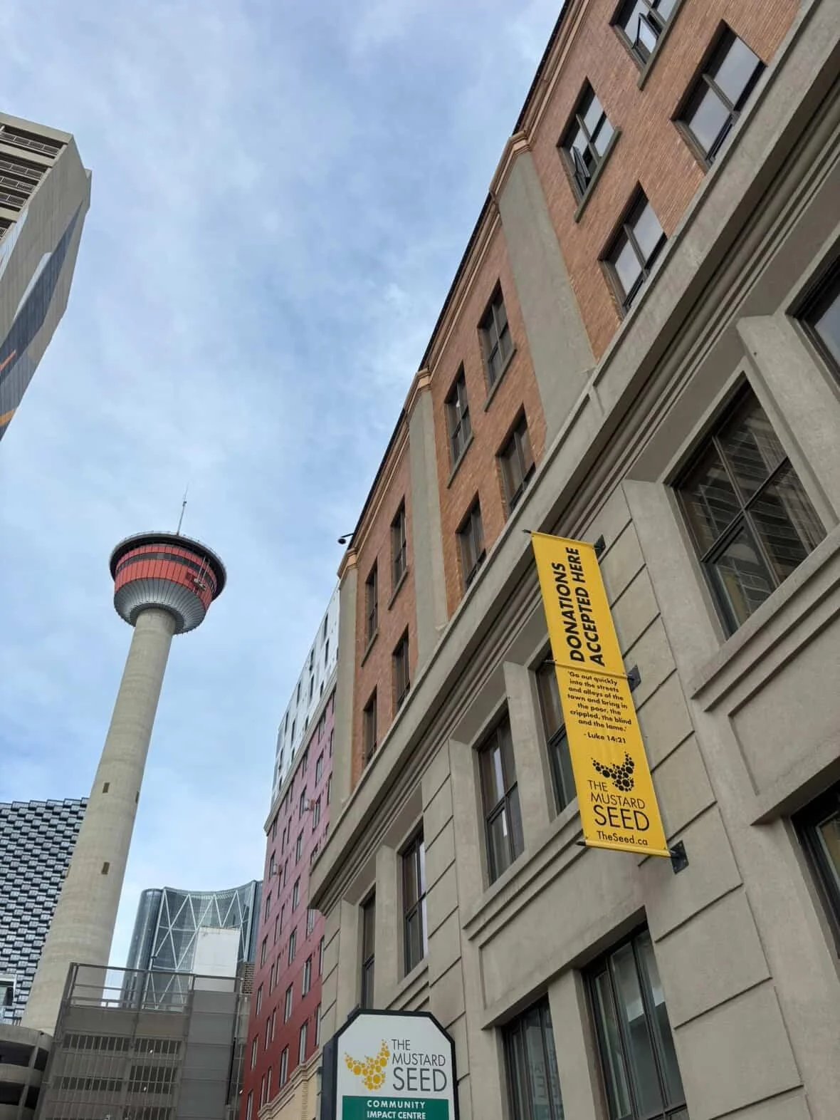 Street view featuring several multi-story buildings with signs for The Mustard Seed and community impact centre, with the Calgary Tower visible in the background.
