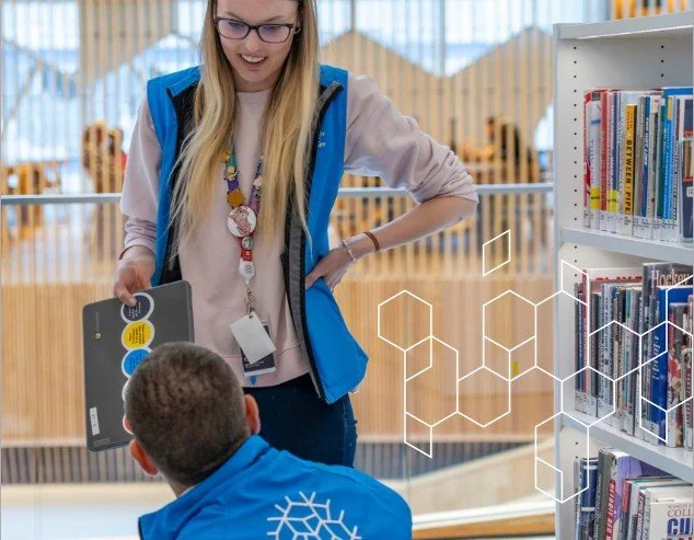 A woman and a boy in a library, with the woman holding a tablet and talking to the boy, who is seated wearing a blue jacket with a white snowflake design. Bookshelves are visible in the background.