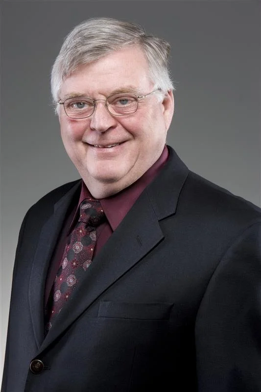 A professional man with gray hair, glasses, wearing a dark suit, maroon shirt, and patterned tie, smiling against a gray background.
