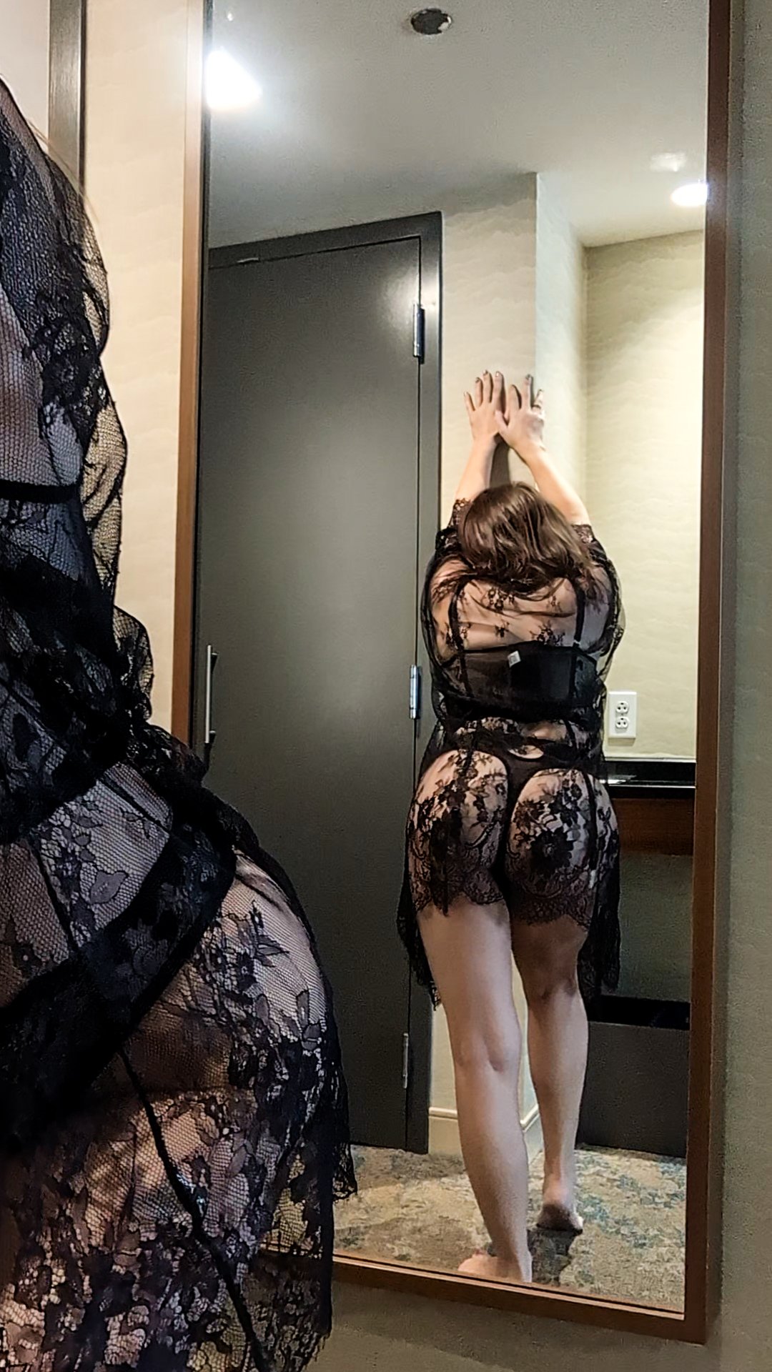 Woman in a black lace robe seen from back, standing in front of a mirror with arms raised above head, wearing matching lace lingerie, in a hotel room.