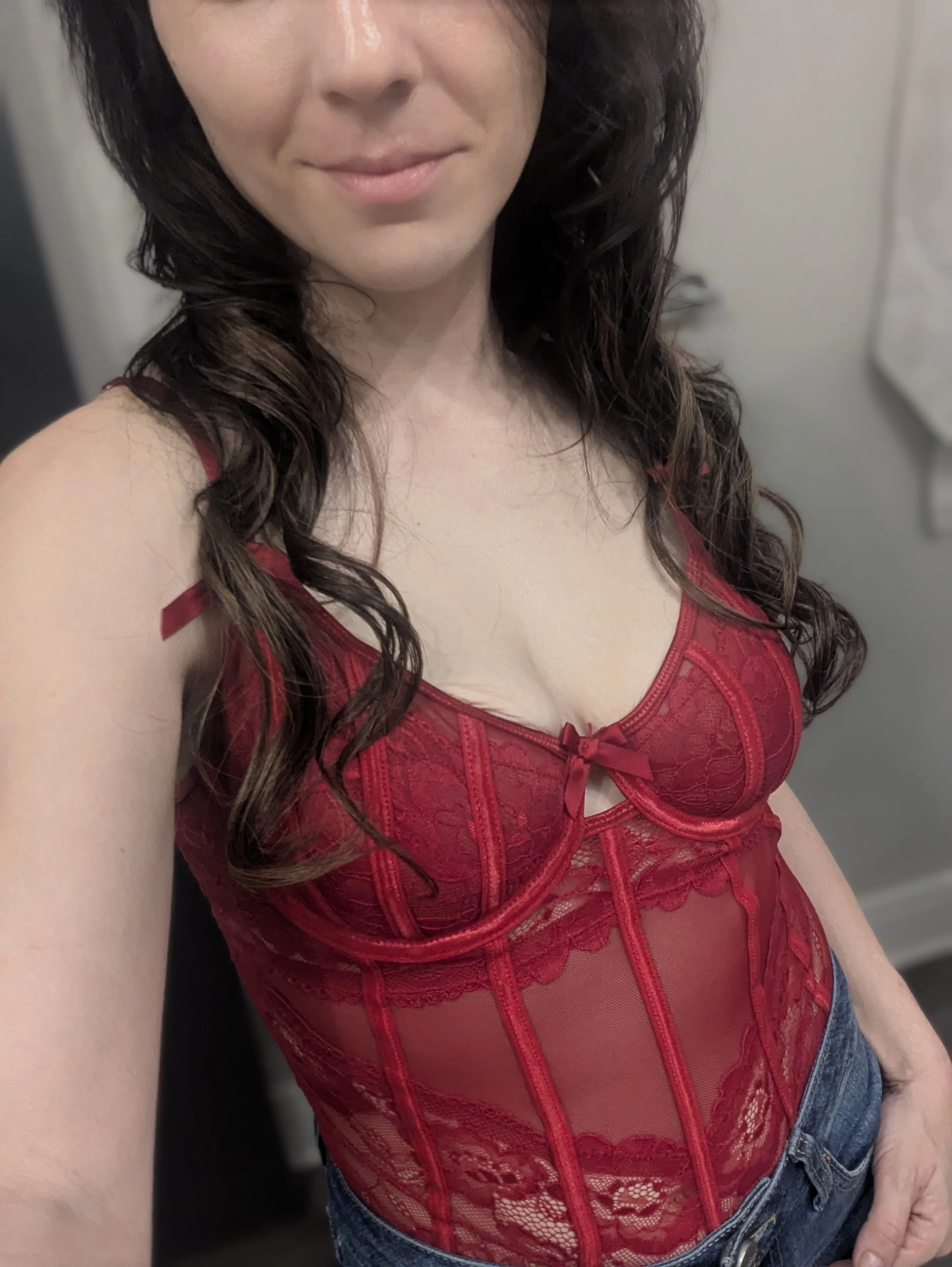 A woman wearing a red lace corset top and blue jeans, taking a selfie in a mirror.
