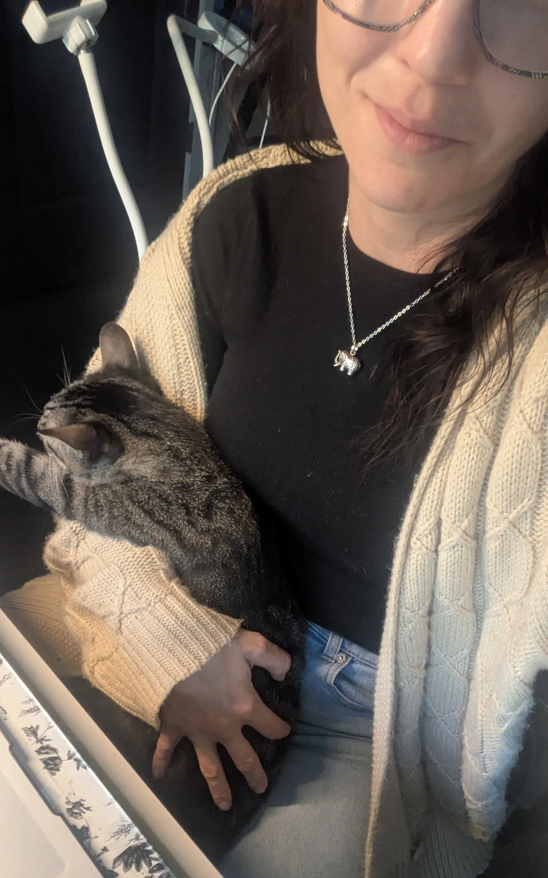 A woman wearing glasses, a black top, a beige knitted cardigan, and a necklace with a rhinoceros pendant sitting with a tabby cat on her lap, holding it with her hand.