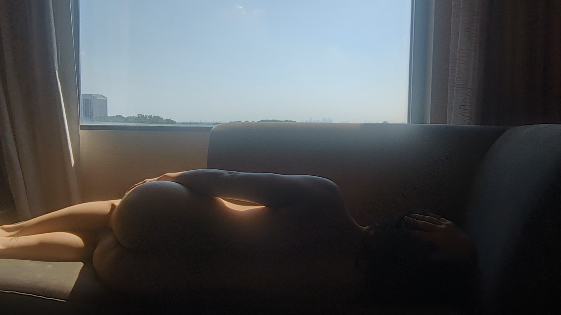 A person lying on a couch near a window, with natural light illuminating their body, and a cityscape visible outside the window.