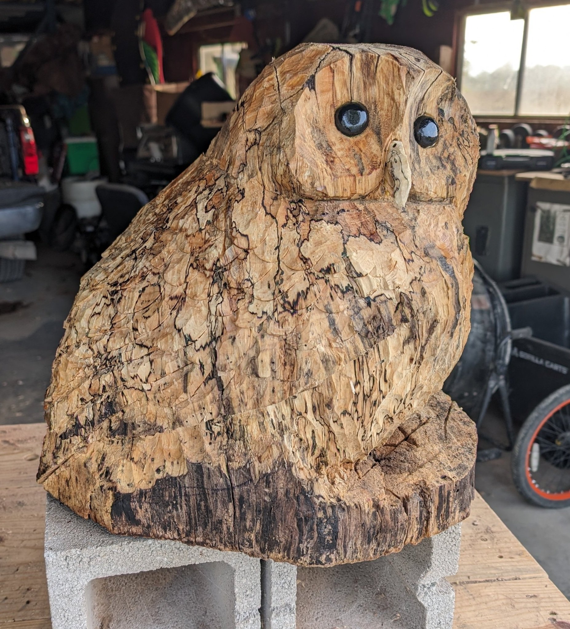 A carved wooden owl sculpture with painted eyes, positioned on a concrete block in a workshop.