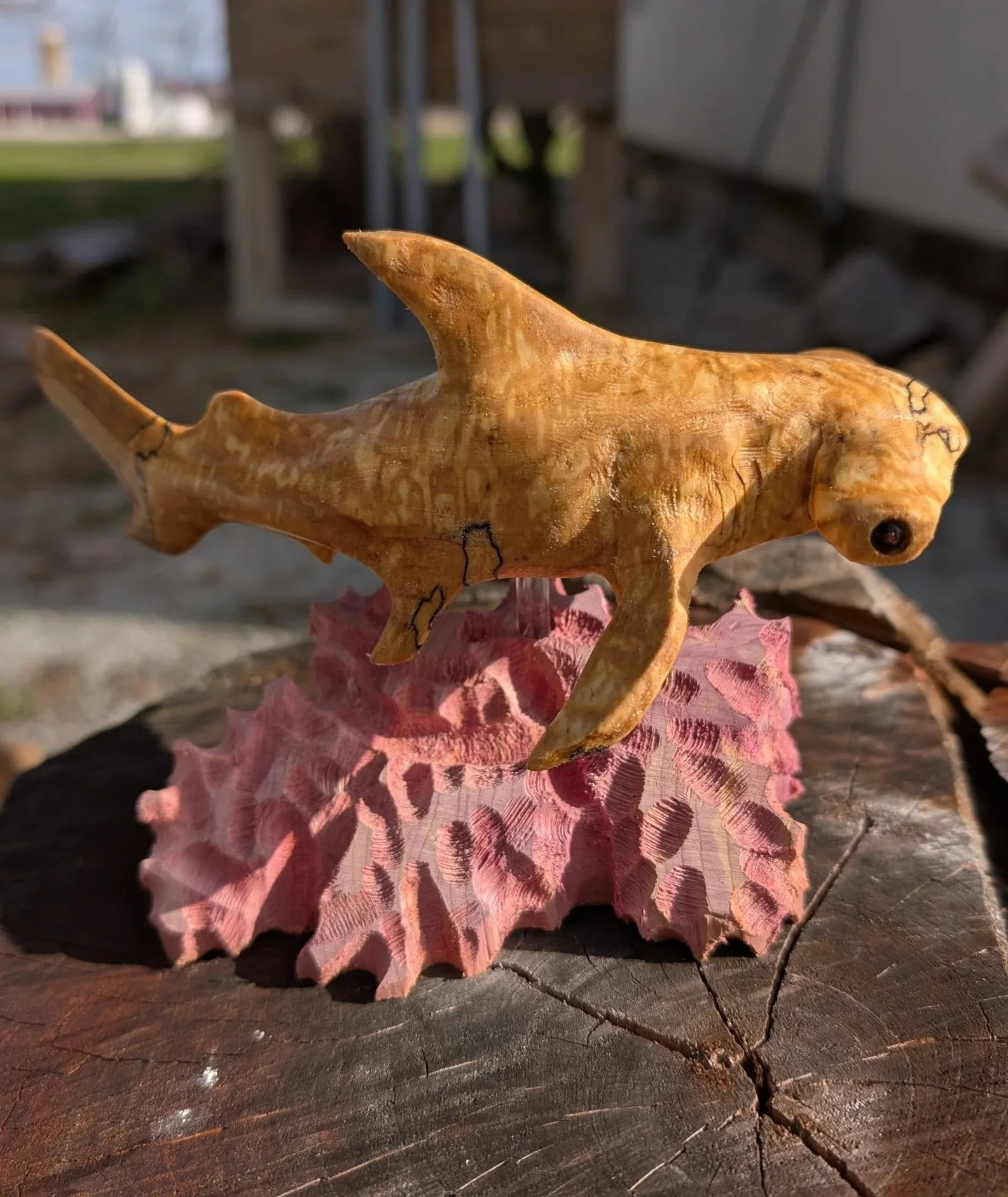 A small carved wooden animal resembling a dinosaur or prehistoric creature, with a horn on its nose, standing on a pink, spiky seashell on a tree stump outdoors.