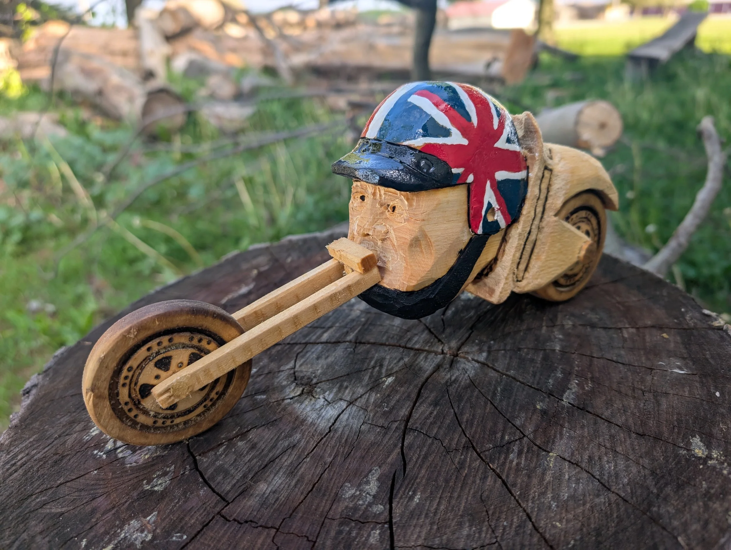 Wooden motorcycle sculpture with a painted helmet featuring the Union Jack flag, placed on a tree stump in a grassy outdoor setting.