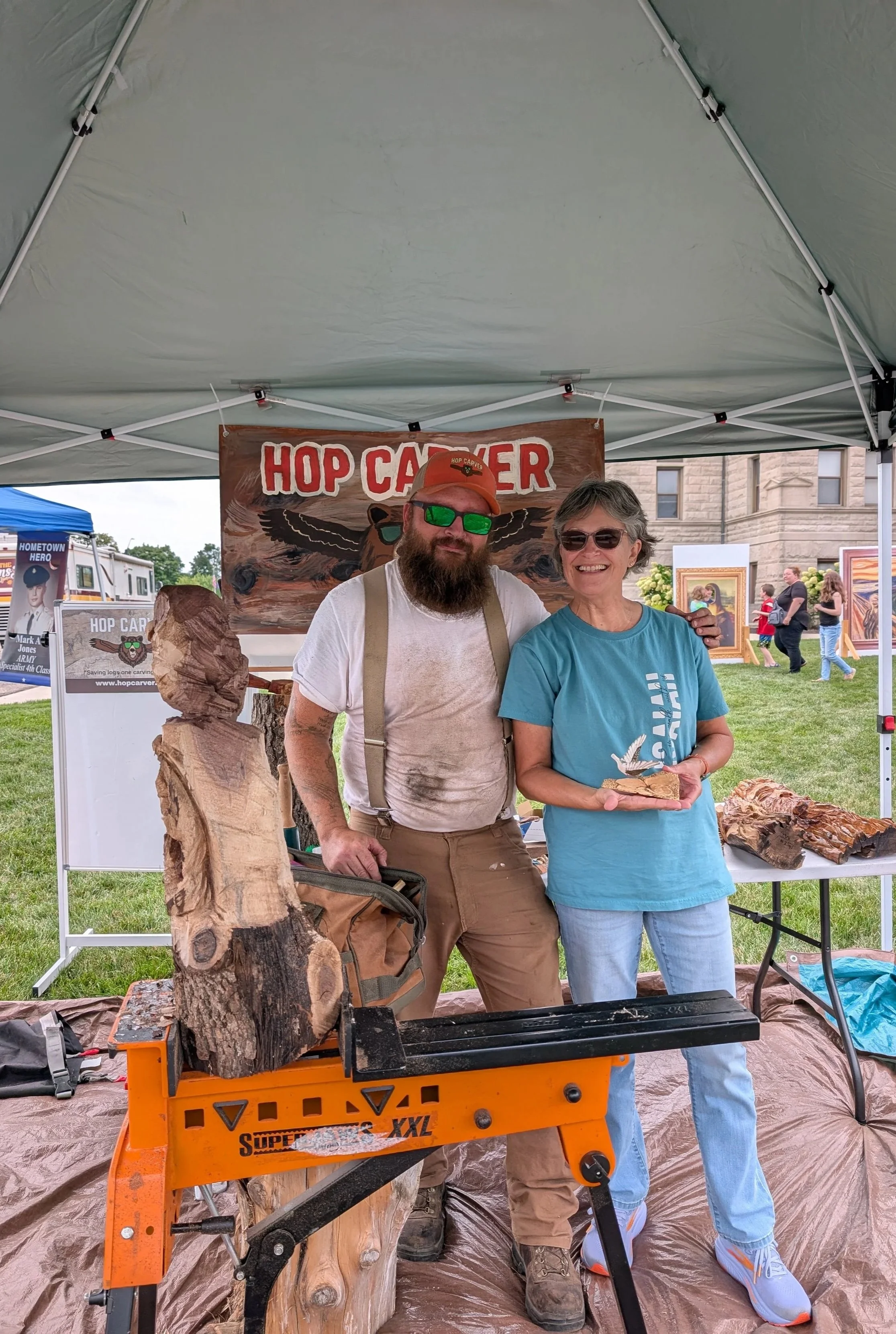 A man with a beard, wearing a cap, sunglasses, and suspenders, standing next to a smiling woman in a blue shirt, holding a wood carving. They are at an outdoor craft fair under a tent with a sign that says 'Hop Carver,' with wood sculptures on display around them.