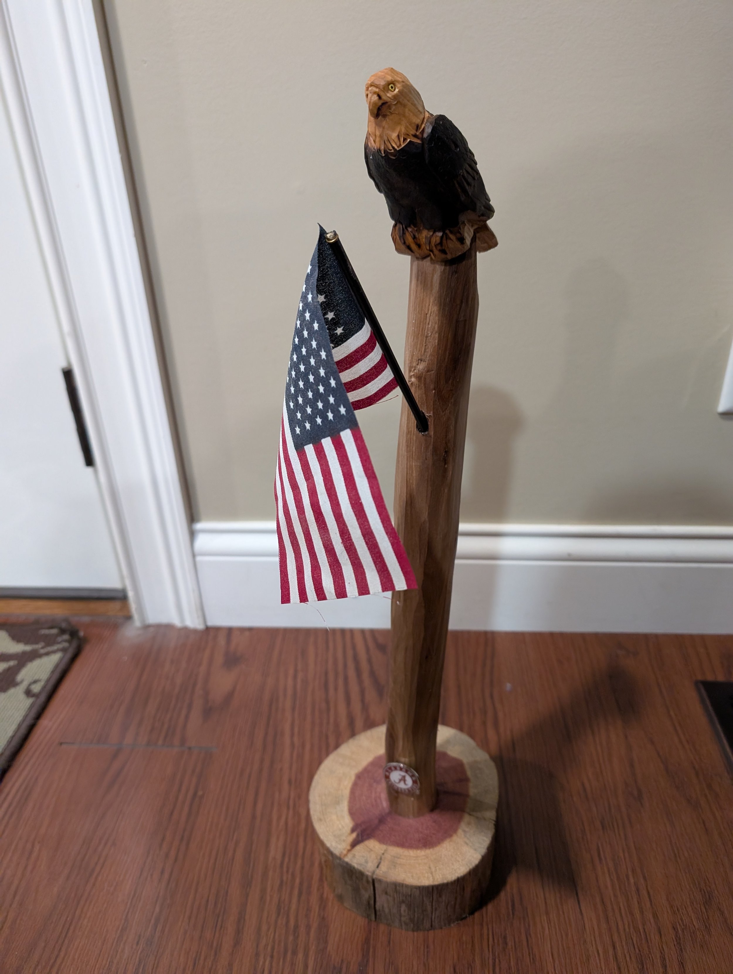 A wooden carved eagle sculpture on a log base with an American flag attached, placed on a hardwood floor.