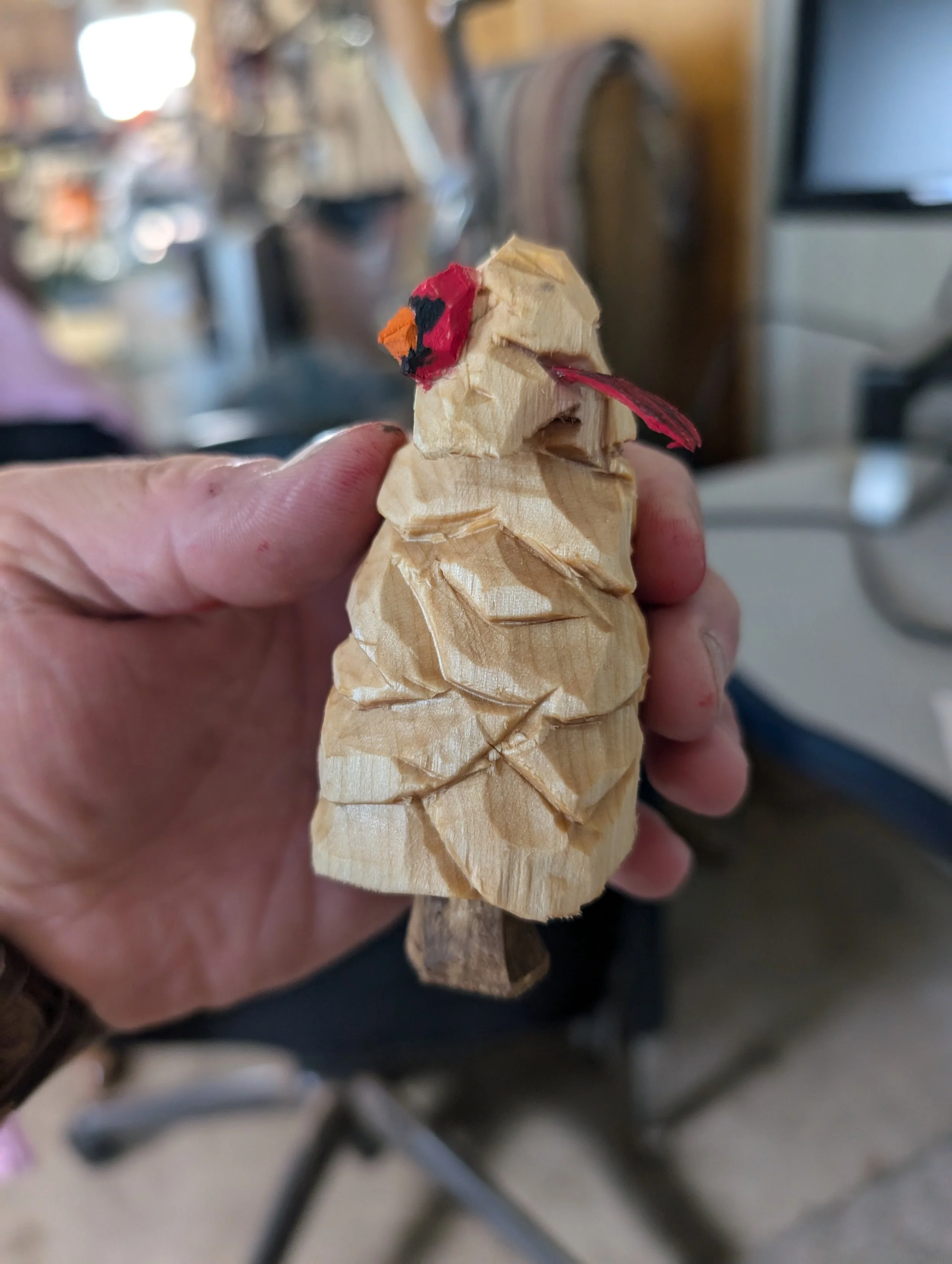 A hand holding a small wooden carved figure of a bird with painted red and black eyes and a red beak. The background shows a blurred room with furniture and a computer.