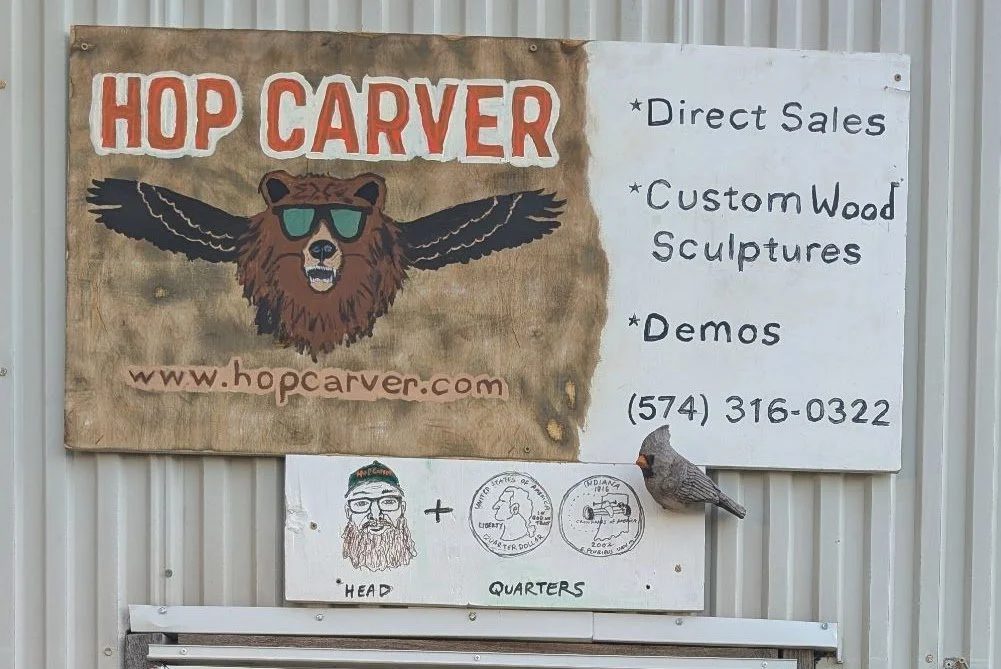 Sign for Hop Carver advertising direct sales, custom wood sculptures, and demos. The sign features a bear wearing sunglasseswith the website hopcarver.com and phone number 574-316-0322. Below the sign, there are illustrations of a bearded man and coins labeled 'quarters', along with a small bird decoration.