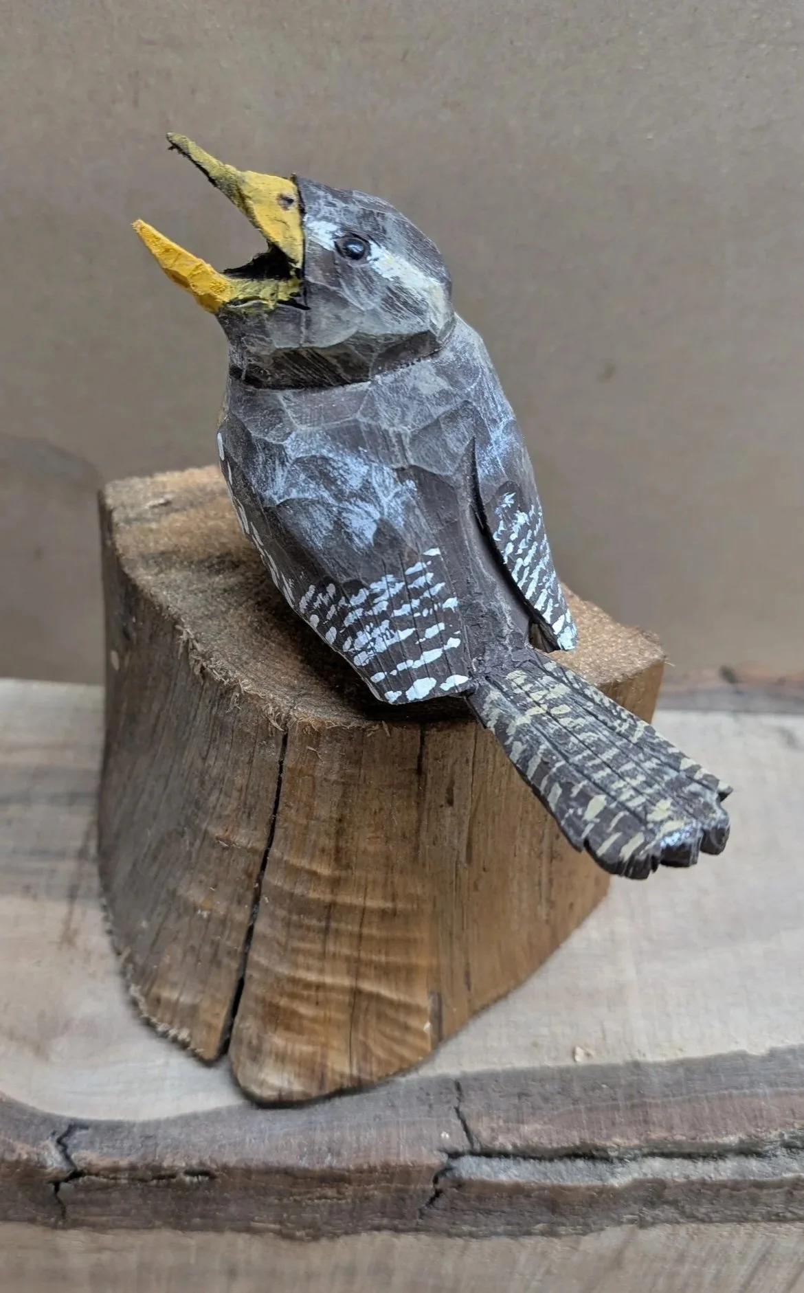 Wooden sculpture of a bird on a log with its beak open, painted in black, gray, white, and yellow, positioned on a wooden surface.