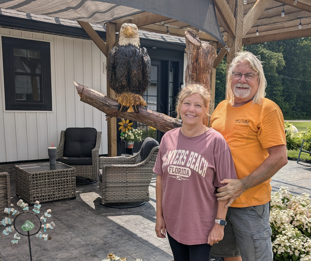A smiling woman in a pink T-shirt and a smiling man in an orange T-shirt stand outside on a patio with a sculpture of a bald eagle perched on a wooden branch behind them. The background shows a white house, outdoor furniture, and greenery.