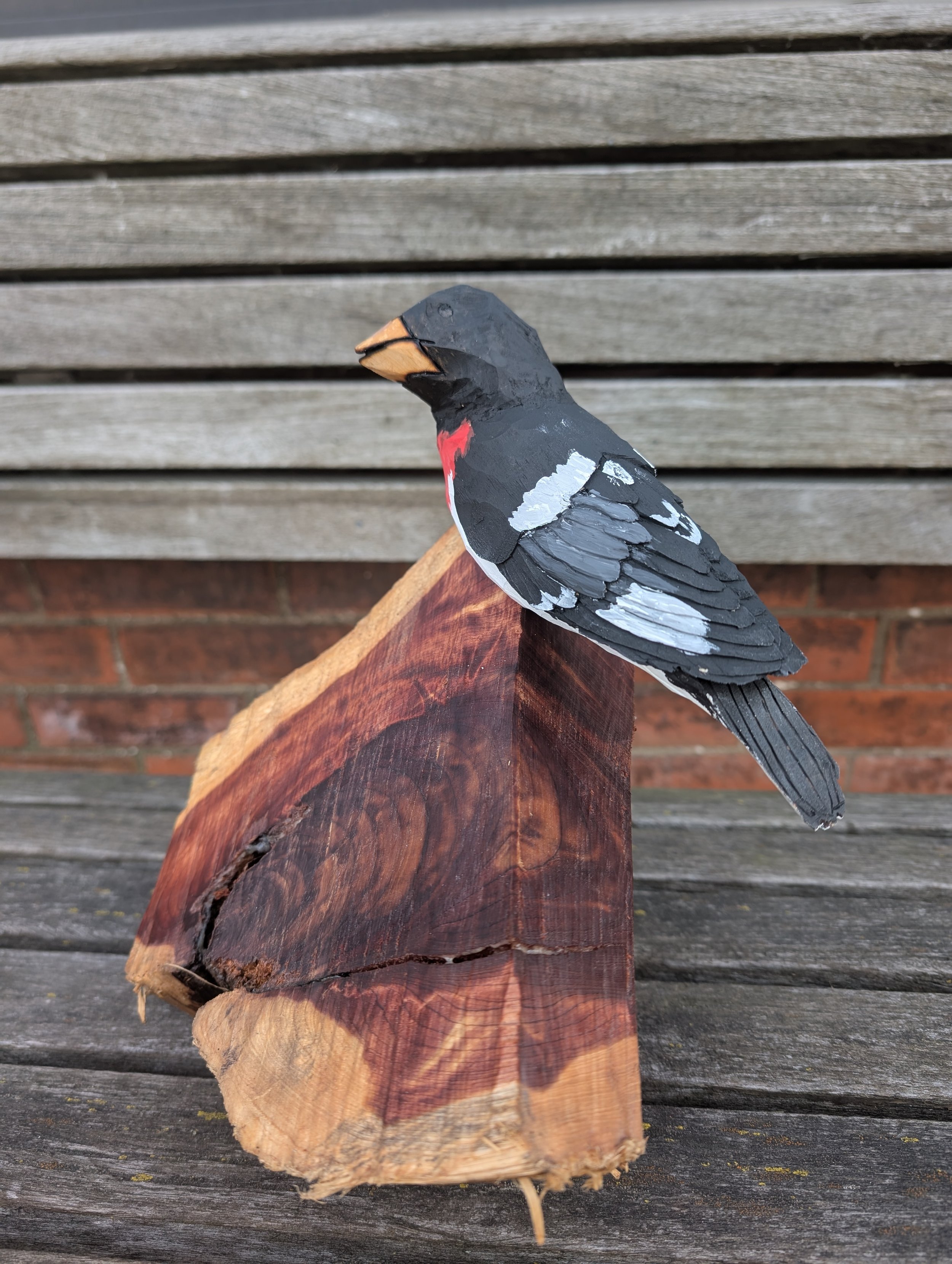 Wooden sculpture of a bird, painted with black, white, gray, red, and orange colors, perched on a natural wood block.