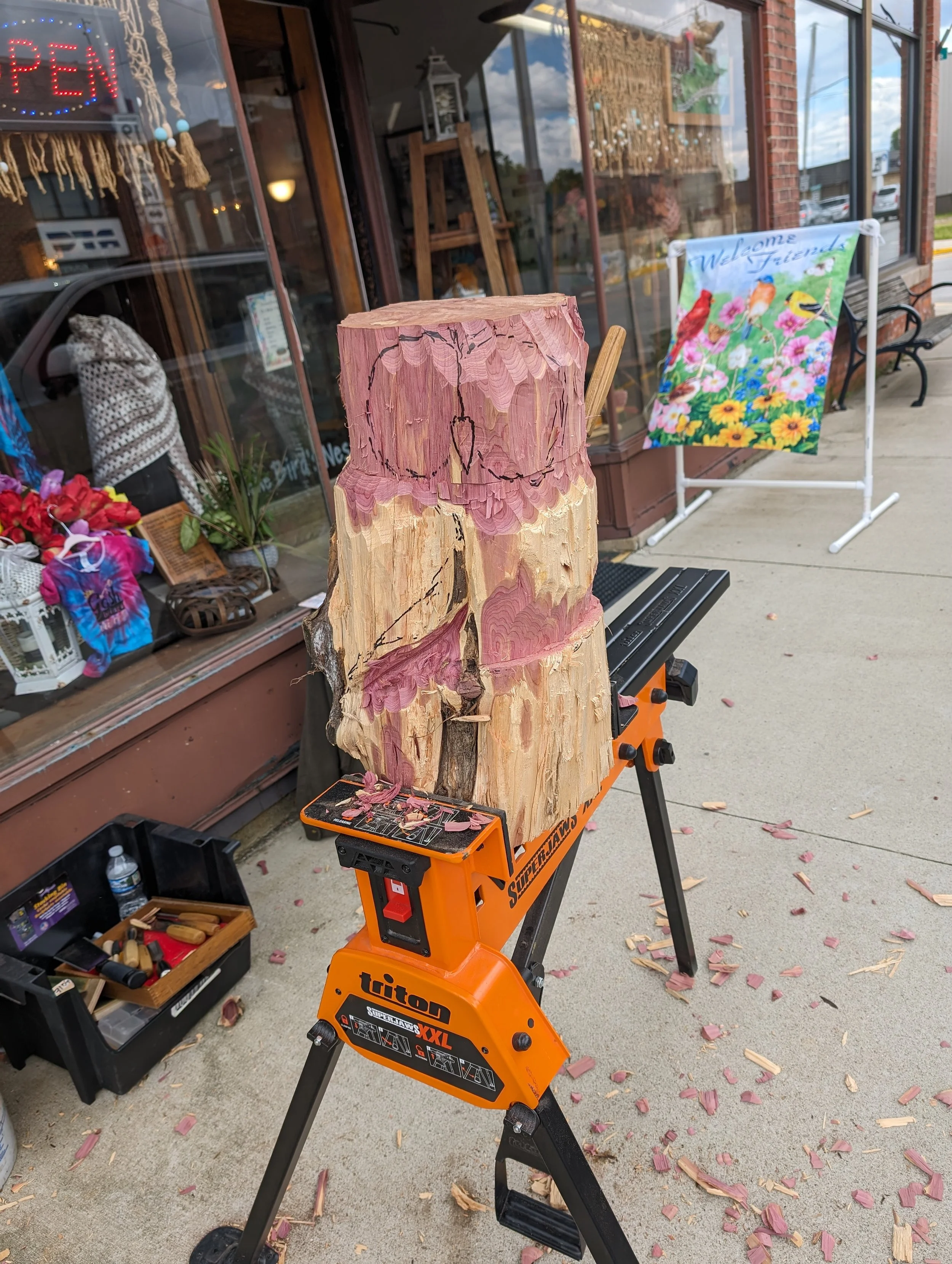 A large wooden log is being carved into a sculpture outside a store. The log is partially shaped and not yet finished. Sawdust is scattered on the ground around the log, which is placed on an orange saw horses. The store window displays various items, including a colorful flower-themed welcome sign and other decorations.