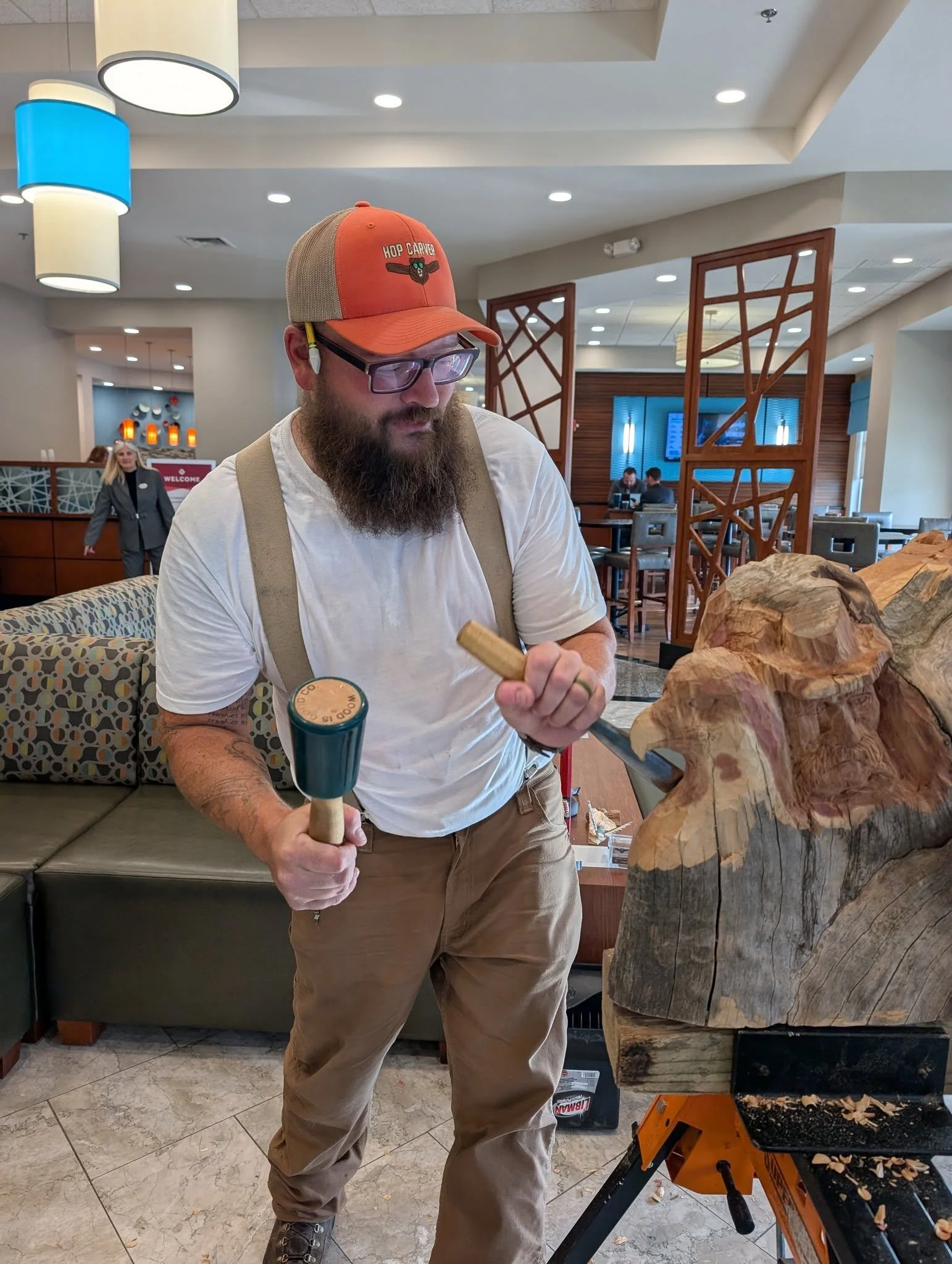 A man with a beard and glasses is carving a wooden sculpture of a face in a public indoor space, possibly a hotel lobby or restaurant. He is wearing a beige and orange cap, a white t-shirt, and khaki pants, and is holding carving tools in both hands.