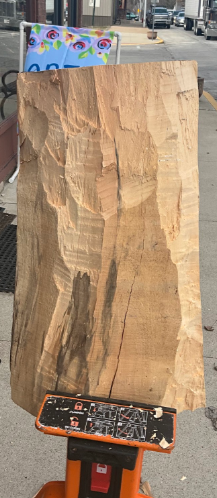 A wooden plank mounted on a wheeled cart on the sidewalk, with a blue box decorated with colorful roses and a colorful mat behind it, in an urban setting.