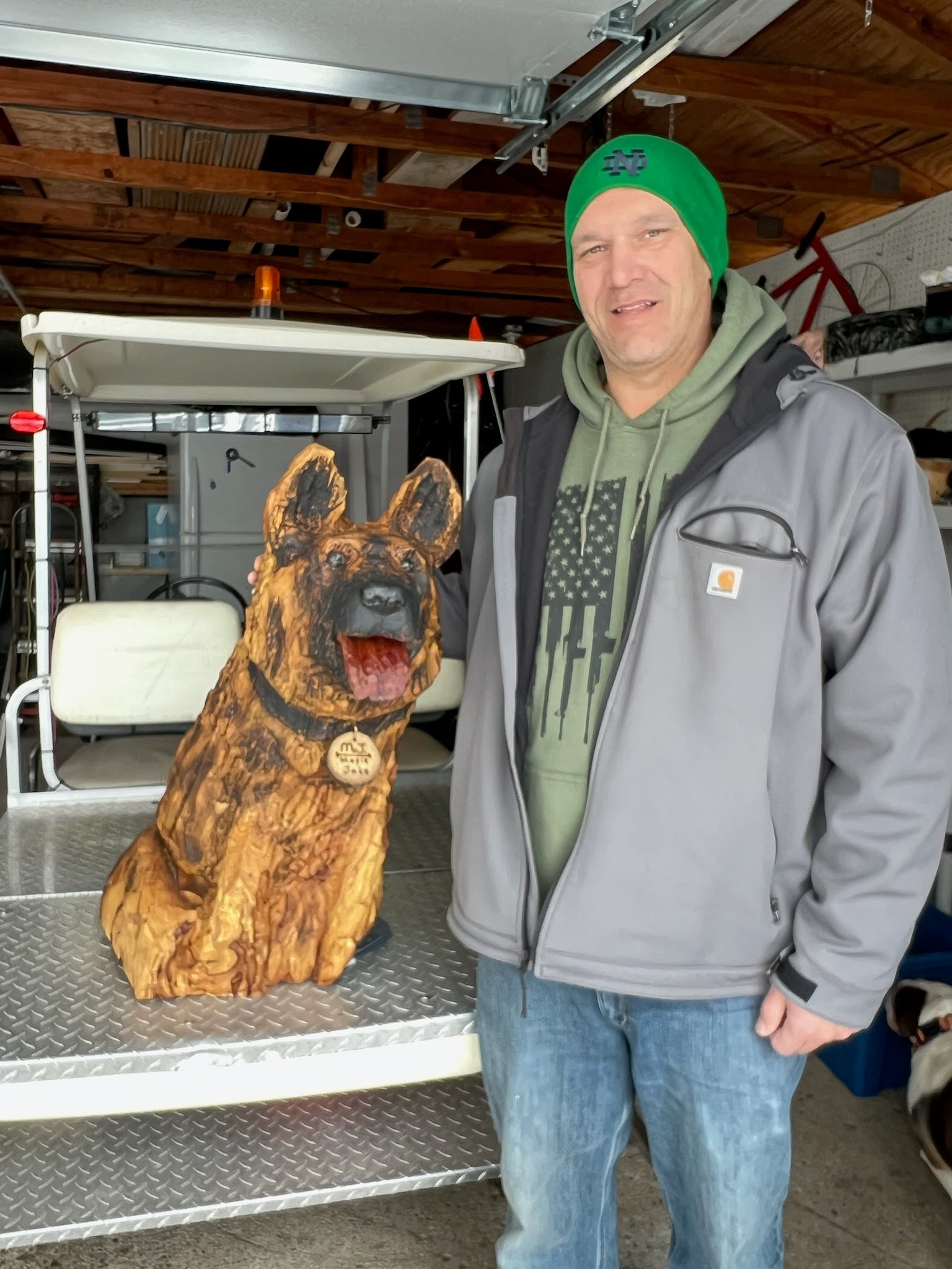A man wearing a green beanie, gray jacket, and green hoodie standing next to a wooden dog sculpture with a carved plaque around its neck, in a garage or workshop.