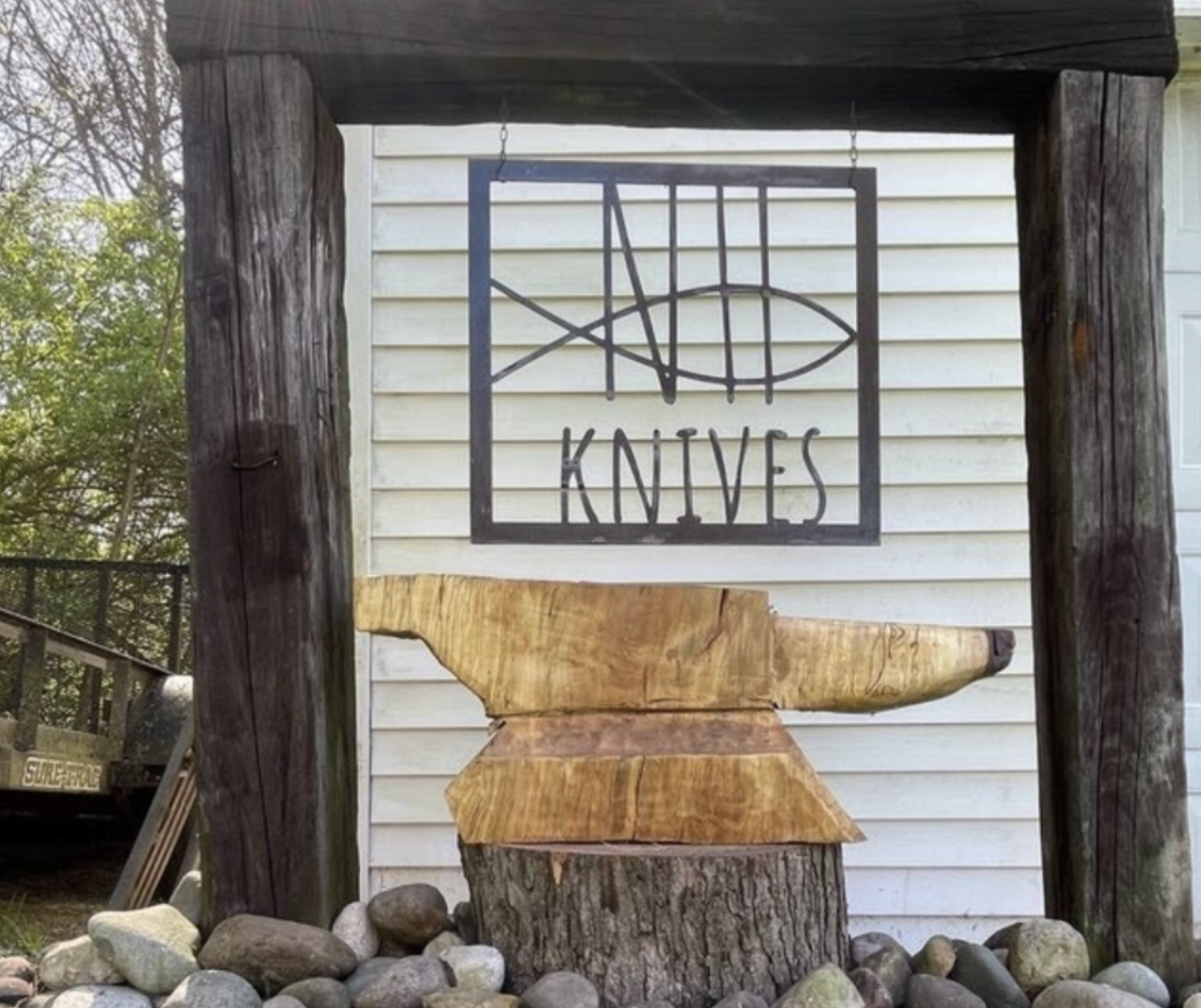Wooden sculpture of a fish and an axe carved from a tree trunk, set within a rustic wooden frame outdoors. Above the sculpture, there's a sign with a fish logo and the words 'KNIVES' underneath.