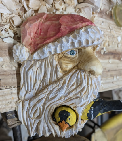 Wood carving of Santa Claus's face with a white beard, a red hat, and a yellow beak, placed on a wooden surface with wood shavings around.