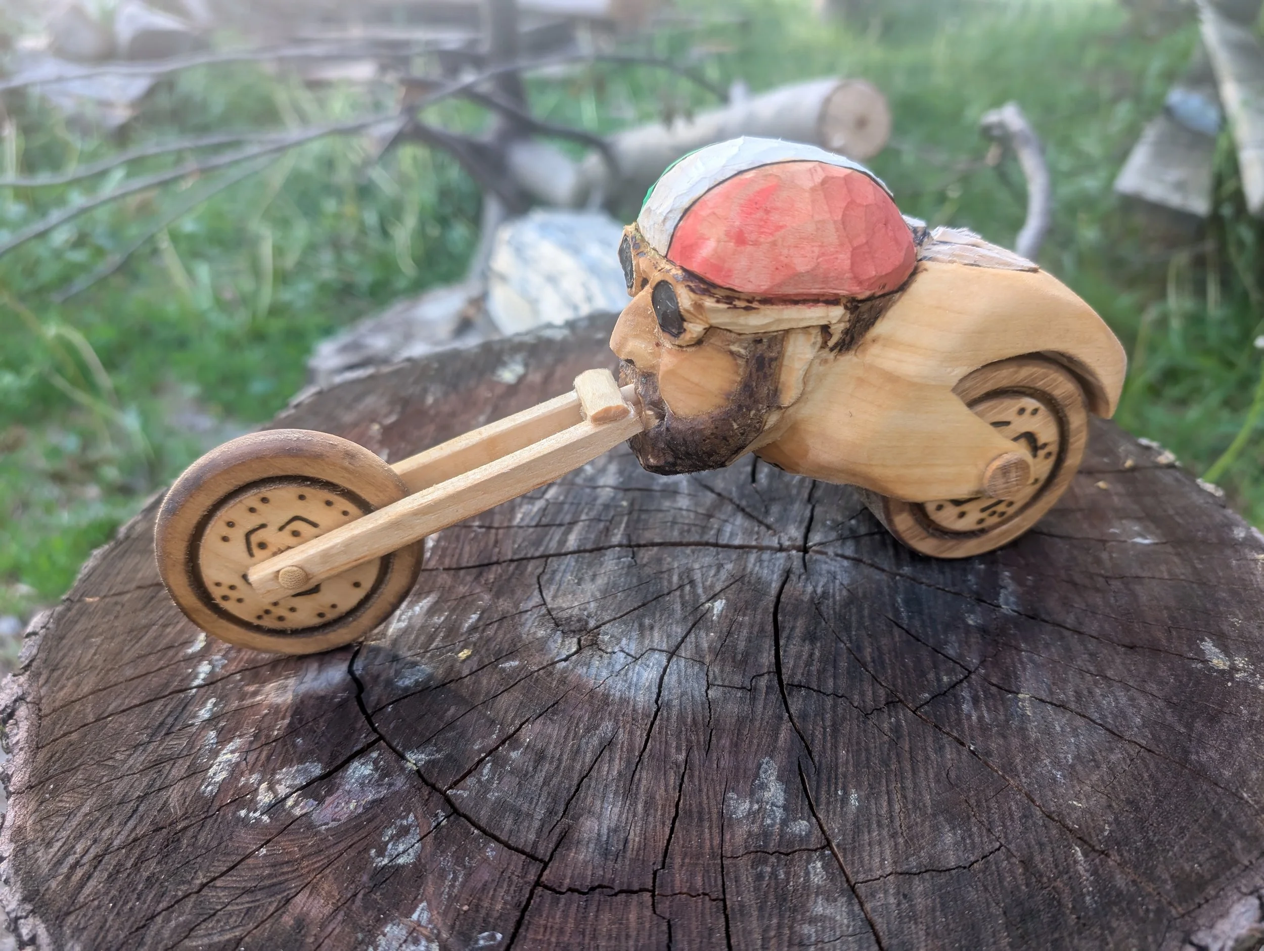 Wooden sculpture of a cyclist with a beard, painted face, and helmet, riding a bicycle, on a tree stump outdoors.