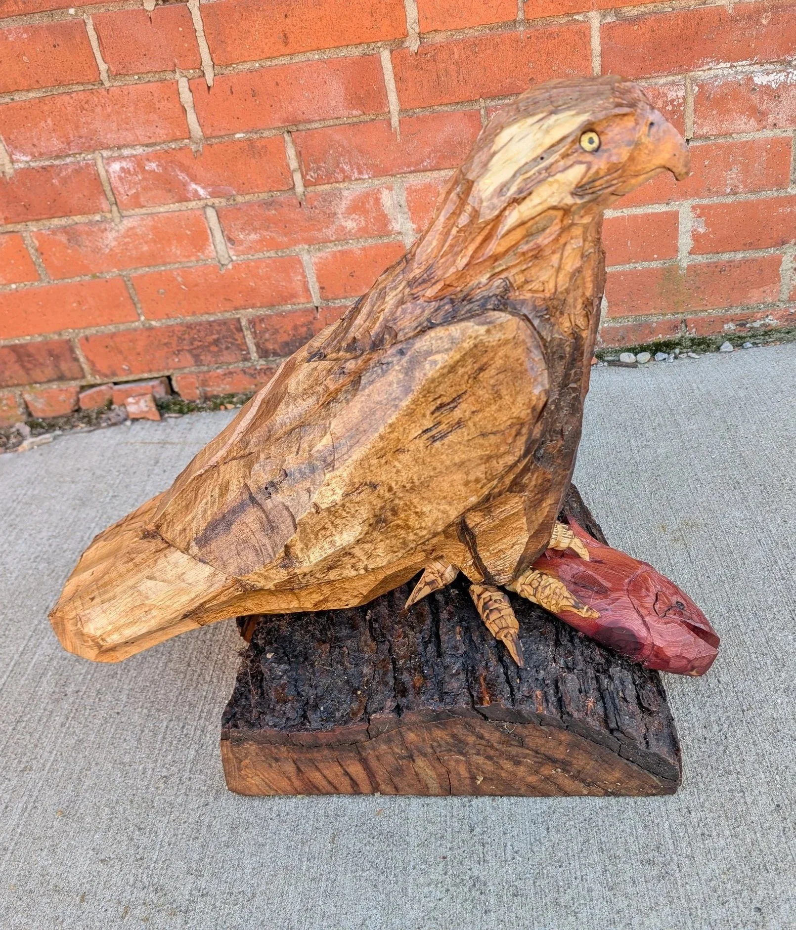 Wooden sculpture of a bird, likely an eagle, perched on a tree stump near a brick wall.