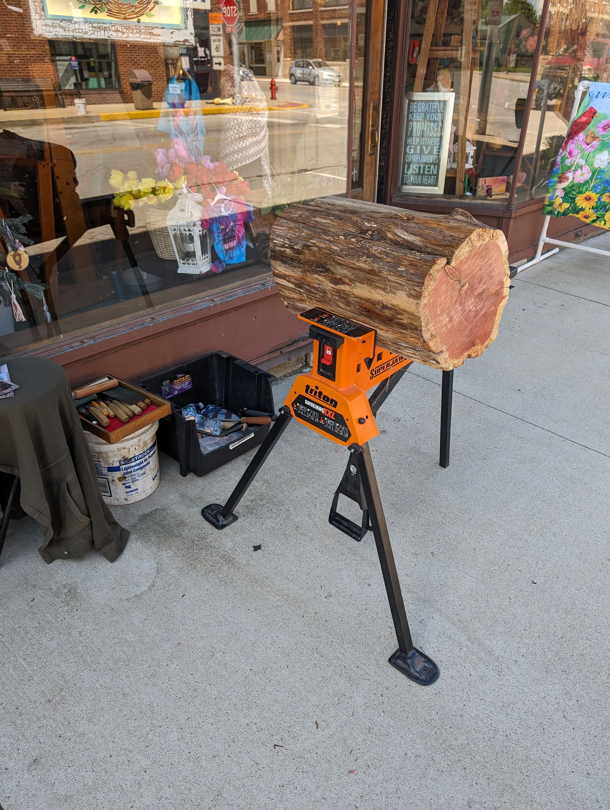 A large log of wood held in place by a saw horse on a sidewalk, with outdoor shop window displays and a reflection of buildings and street in the glass.