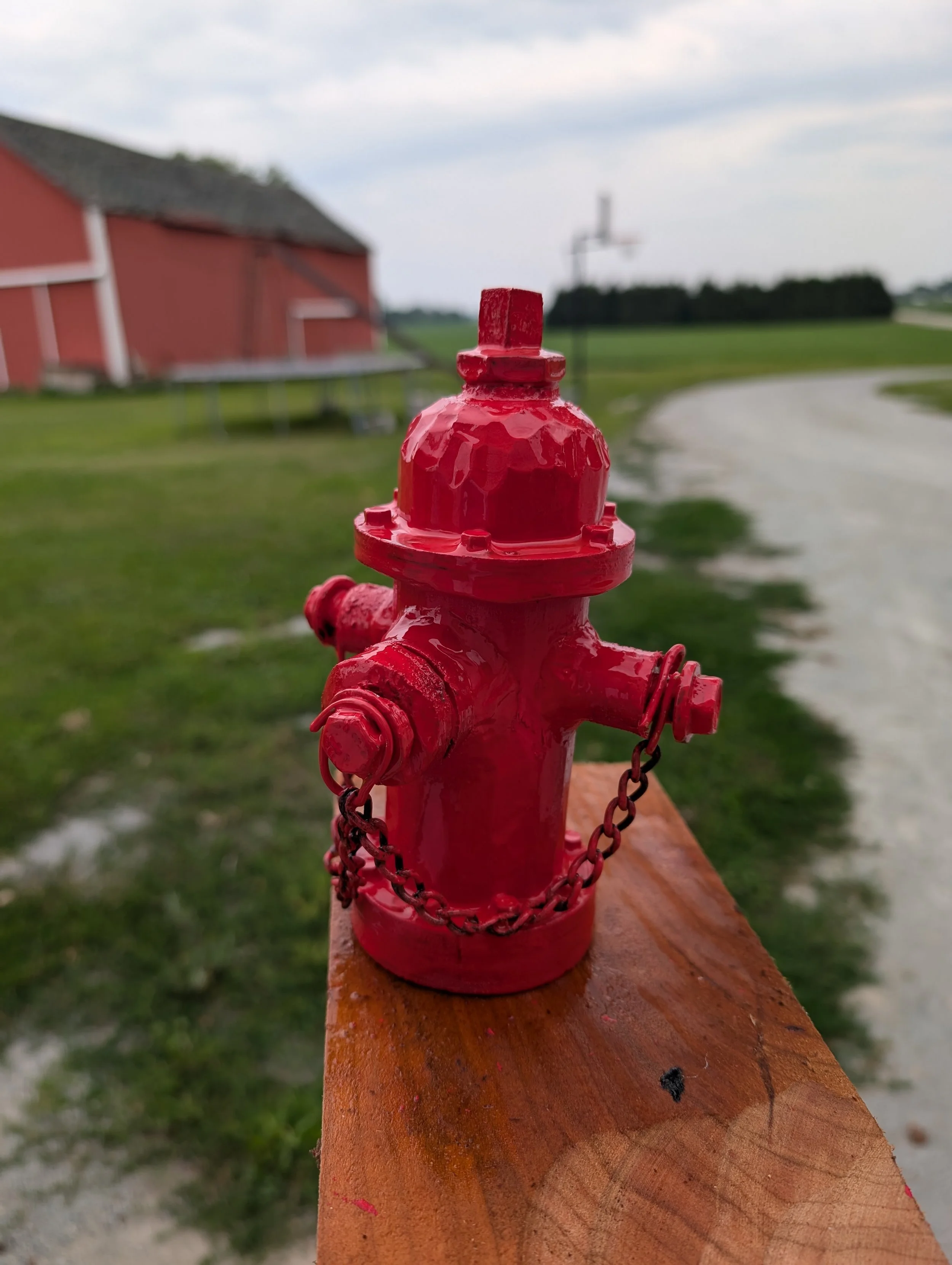 Red fire hydrant on a wooden post outdoors with a red barn and a dirt path in the background.