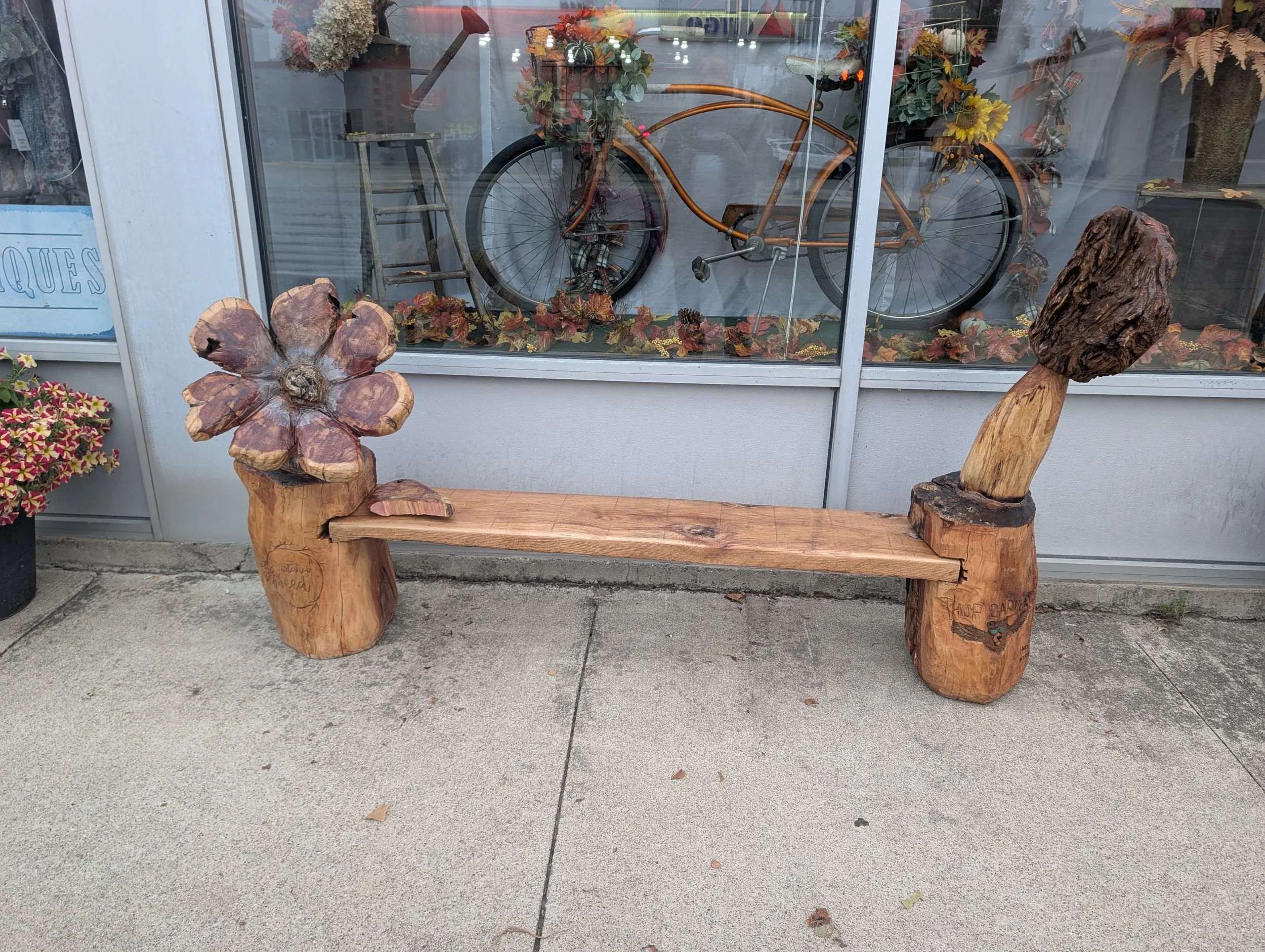 Rustic wooden bench with a flower carved from wood on the left side and a head made from a piece of wood on a stand on the right side, placed outside in front of a shop window displaying a bicycle decorated with flowers and autumn leaves.
