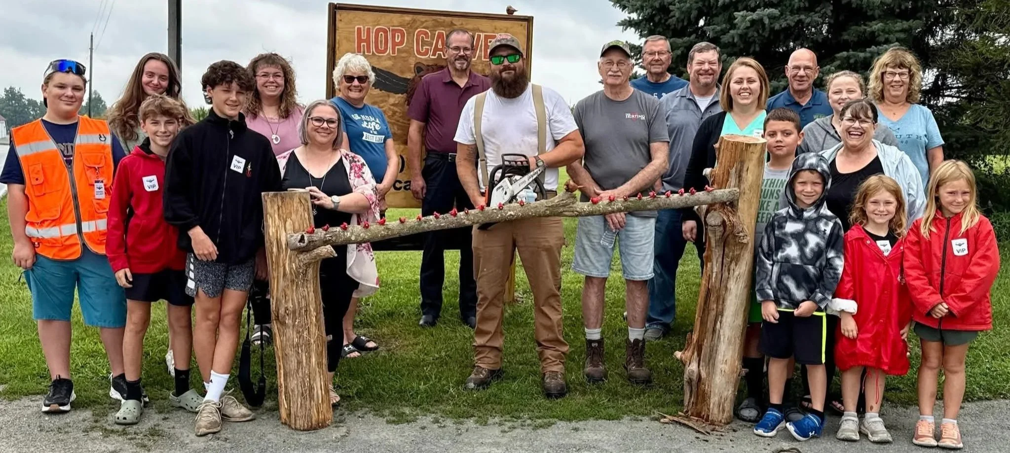 Group of people at a tree-planting event, with children and adults standing behind a fallen tree on supports, in front of a sign that reads "HOP CAVE." The group is smiling and outdoors on a cloudy day.