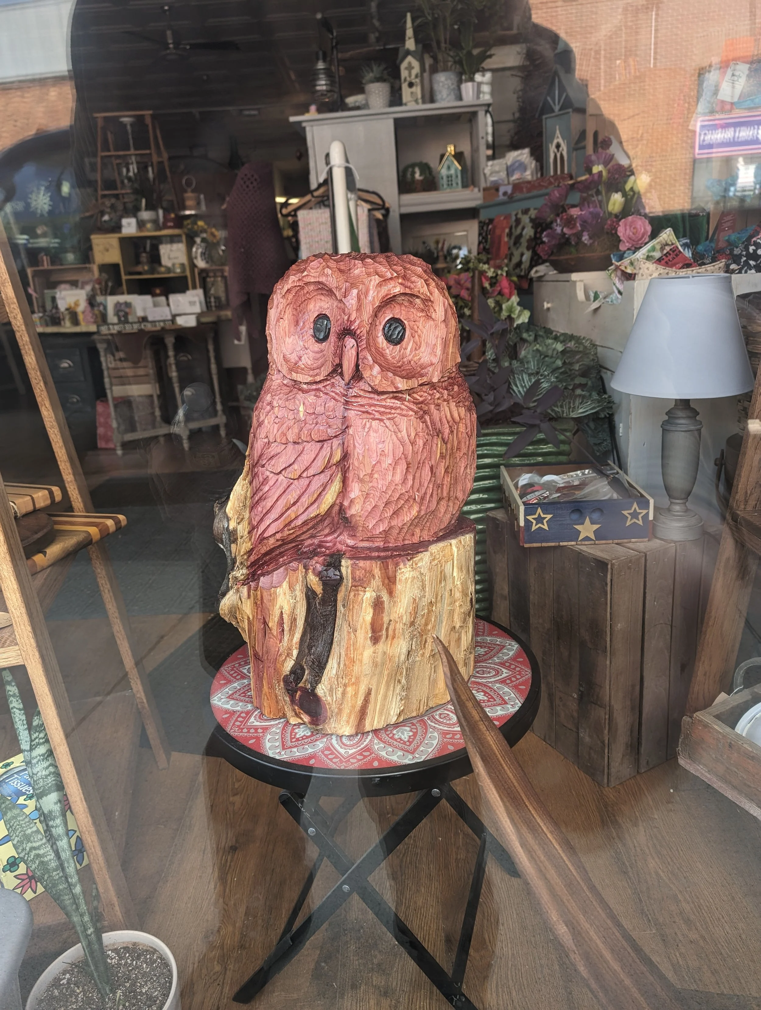 Decorative carved owl sculpture on a small round table inside a store, viewed through a glass window.