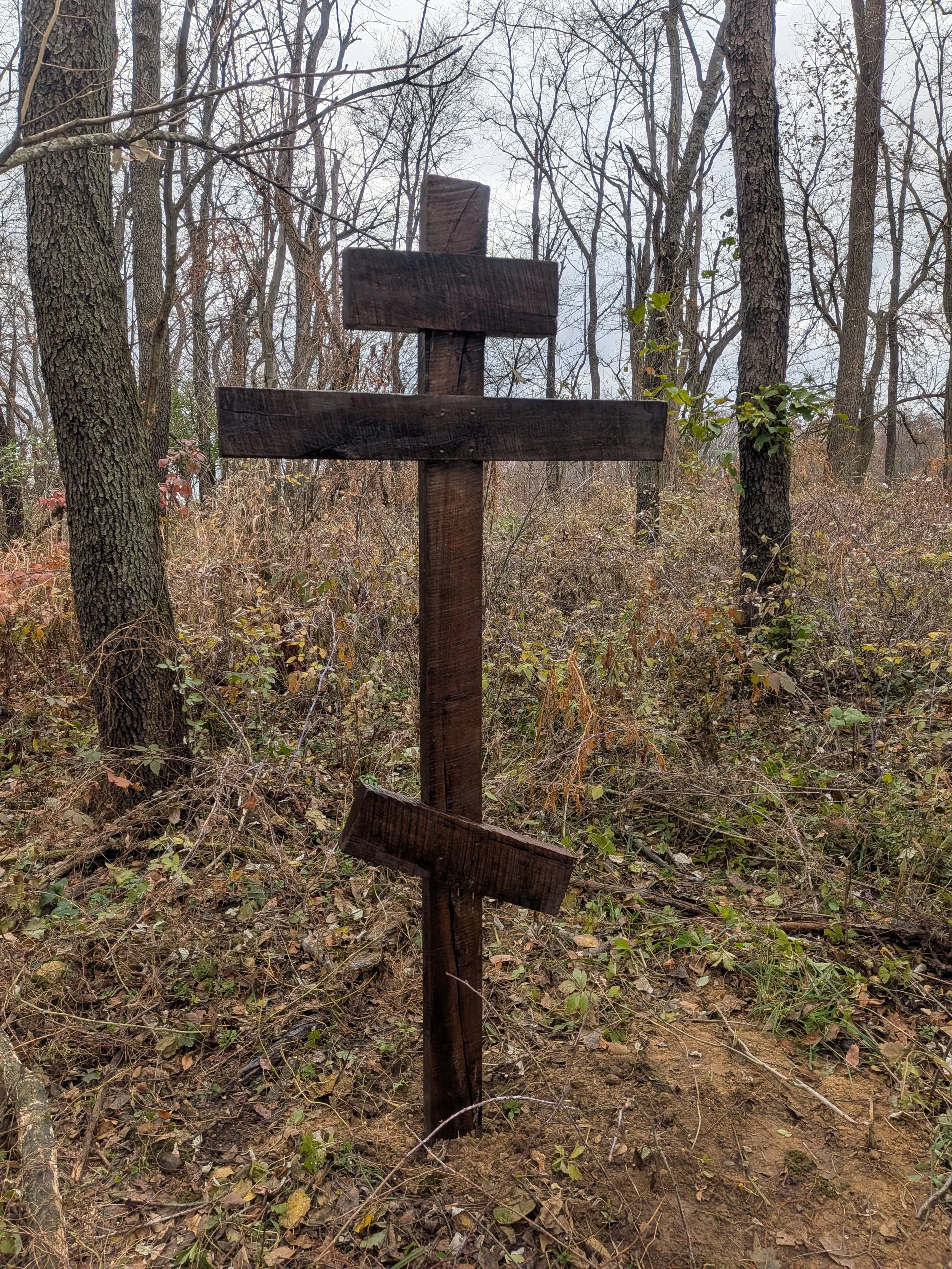 A wooden cross with three horizontal slats, standing in a wooded area with leafless trees and sparse vegetation.