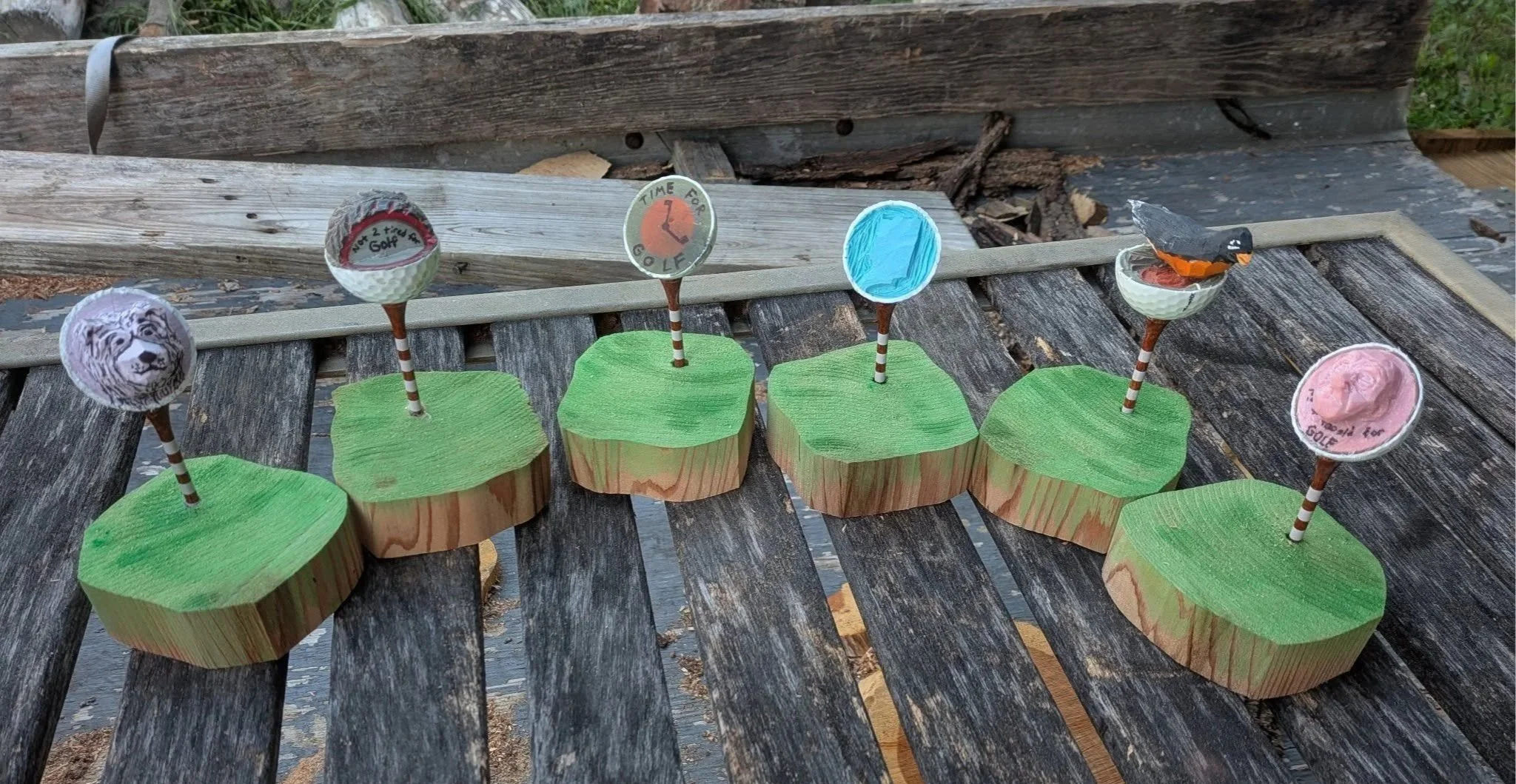 Wooden golf ball markers decorated with animal images, golf-related stickers, and painted designs, placed on small wooden bases painted green, arranged on a weathered wooden surface.
