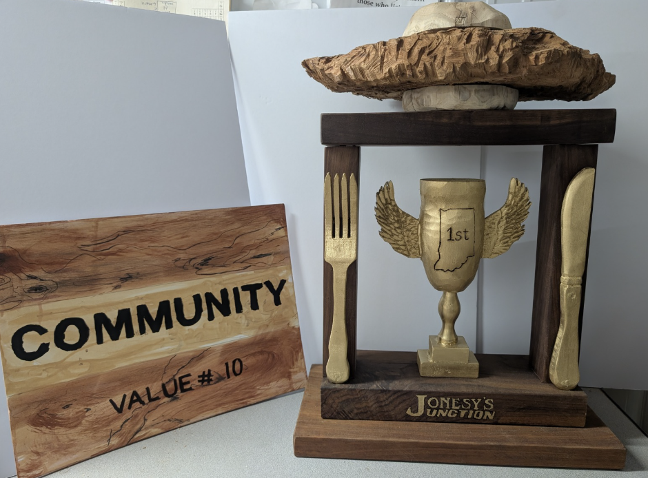 Decorative wooden trophy with a gold cup and two wings, labeled '1st', on a wooden stand. A large wooden fork and knife are on either side of the trophy, all on a wooden table. A sign with the word 'COMMUNITY' and 'VALUE #10' is next to the display.
