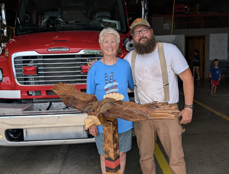 A woman and a man stand together inside a fire station, posing with a wooden eagle sculpture. The woman is wearing a blue shirt with a hand making a peace sign, and the man is wearing a white T-shirt, suspenders, and a cap. Behind them is a red fire truck