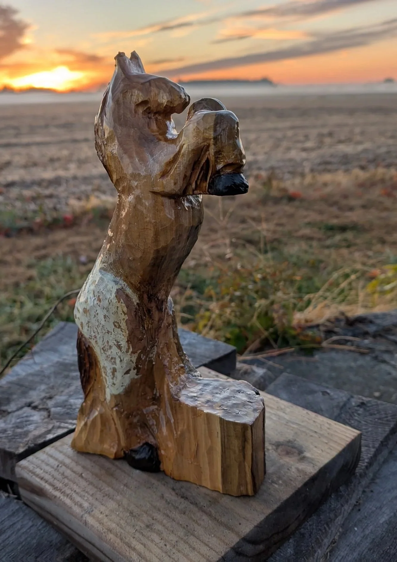 A wooden sculpture resembling a wolf or fox howling at sunset, set outdoors on a wooden base with a sunset sky in the background.