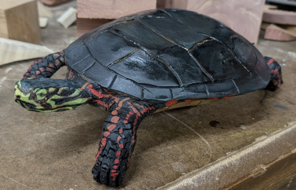 A realistic model of a turtle with a black shell, green and yellow head, and red and black limbs, placed on a wooden surface.