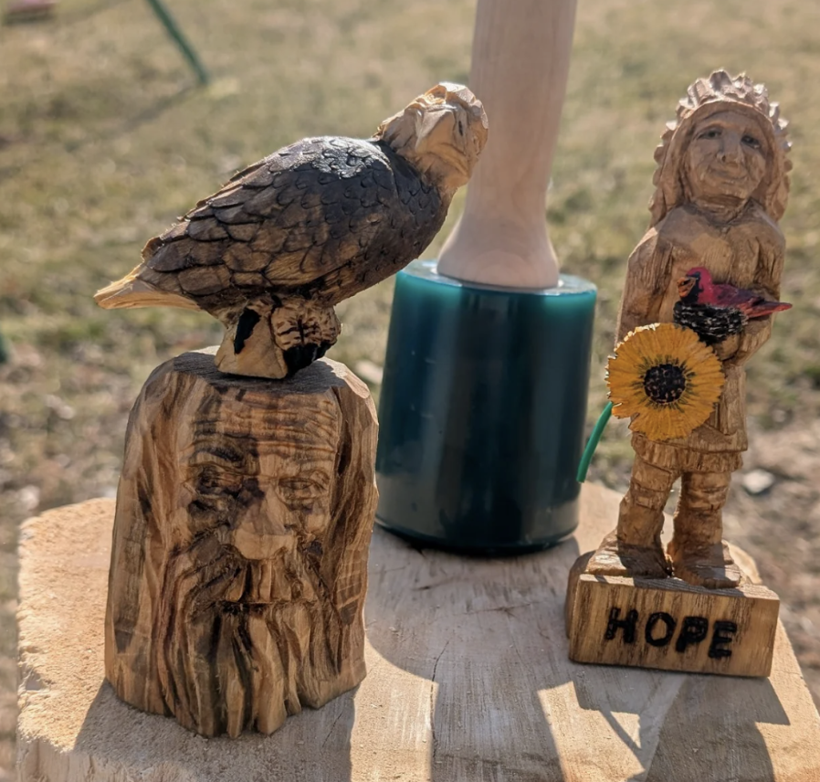 Wooden carvings of a bearded man's face, a bird, a native child holding a sunflower with the word "HOPE" on the base, and a lamp in the background on a wooden surface.