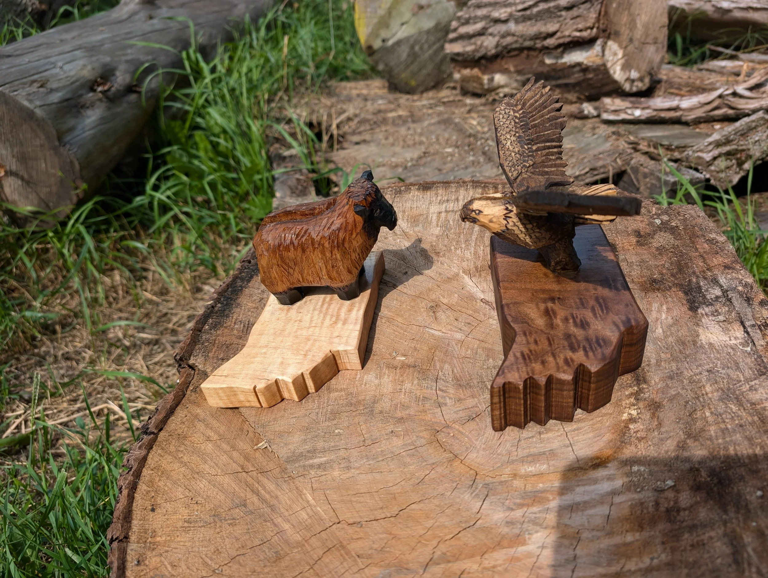 Two handcrafted wooden animal figurines, a bison and a bird of prey, are displayed on wooden bases on a large tree stump outdoors.