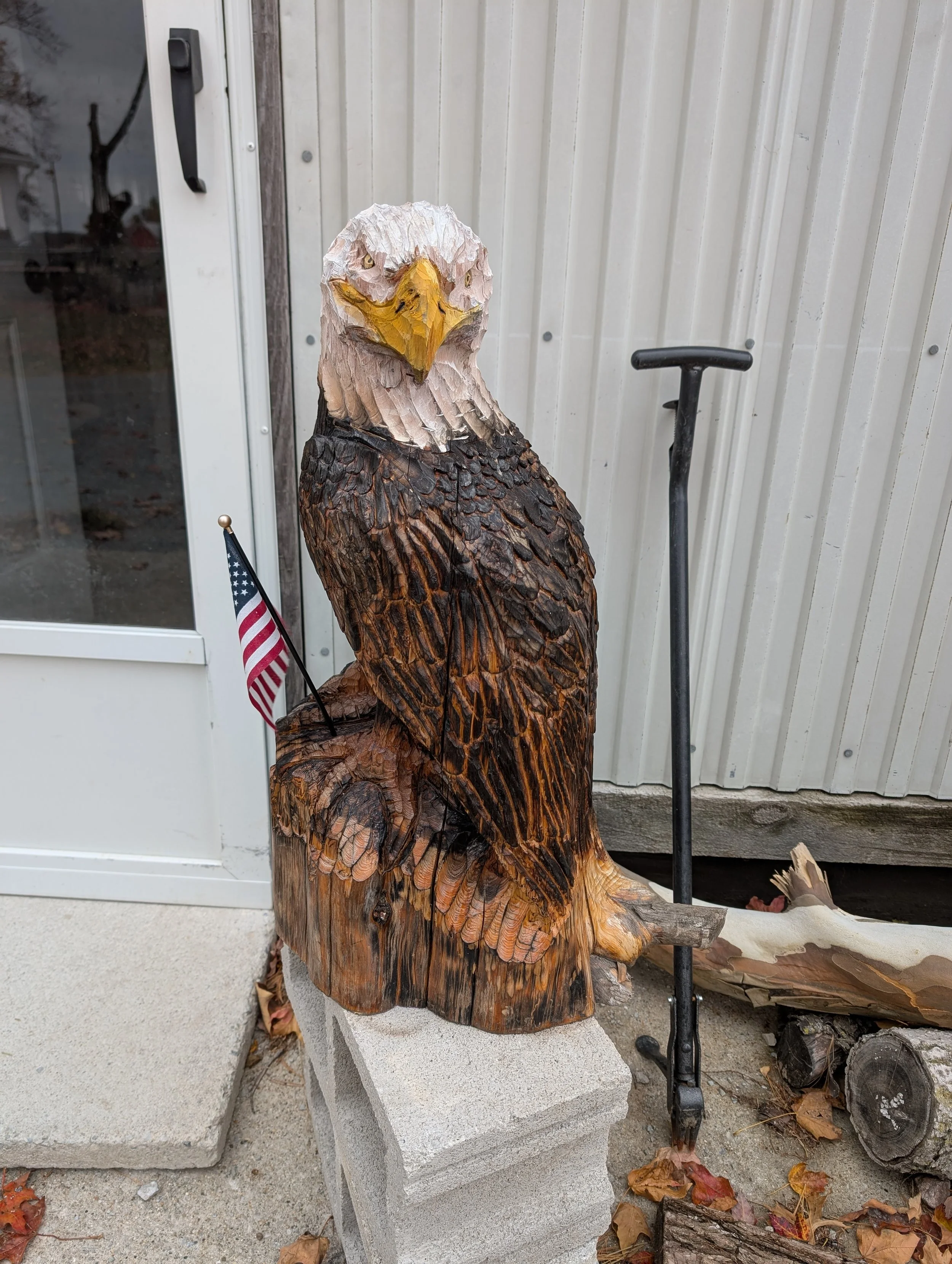 Wood carved eagle sculpture with American flag and a tool leaning against a building exterior.
