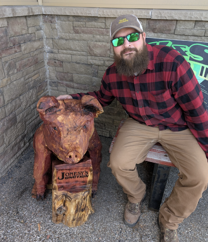 A man with a beard wearing sunglasses, a baseball cap, and a red and black flannel shirt sitting on a bench next to a carved wooden pig statue.